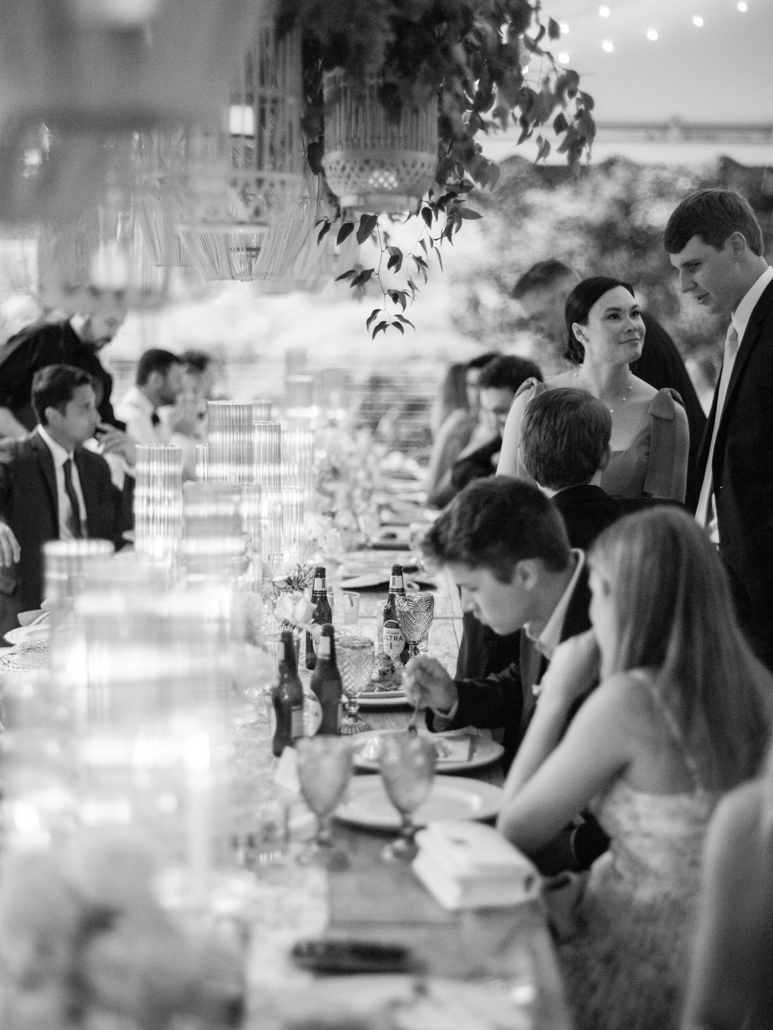 A black and white photo of a formal gathering at a long table with people dining and socializing, with hanging decorations above.