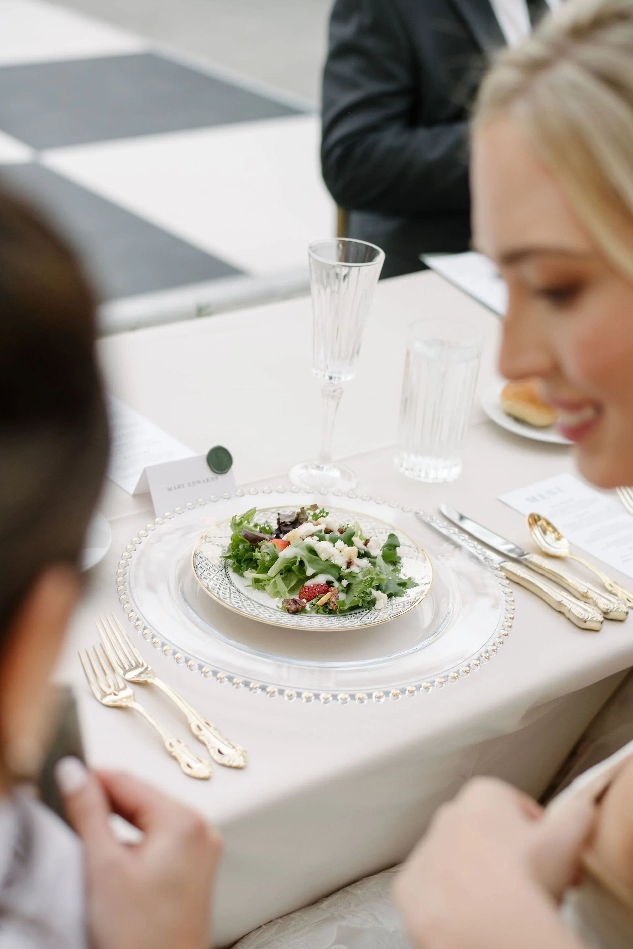 Elegant dining table with a white tablecloth, gold utensils, a floral salad on a patterned plate, a water glass, a champagne flute, and a place card.