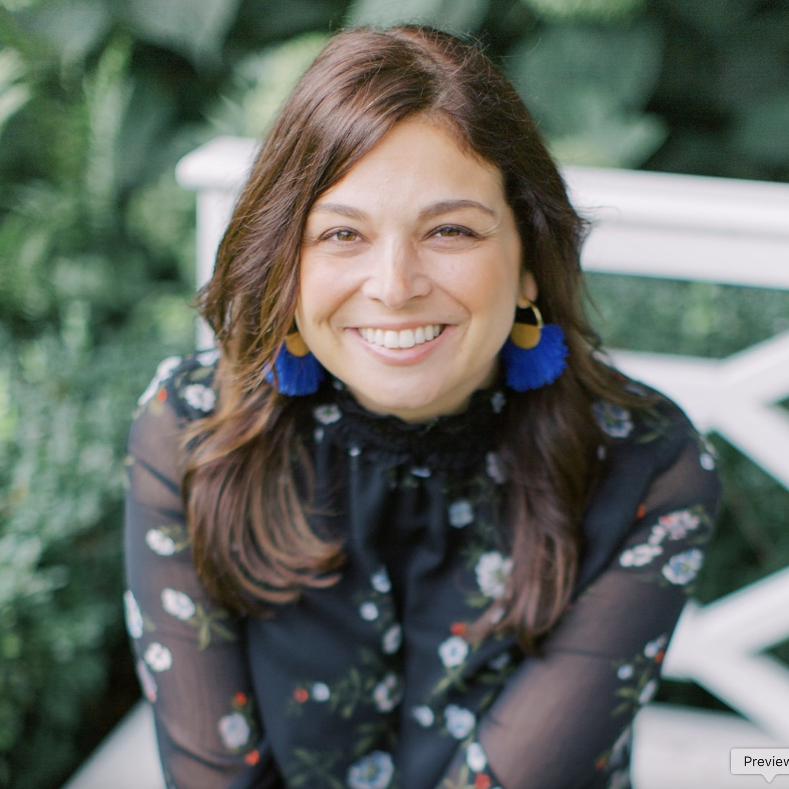 Close-up of a smiling woman with long dark hair, resting her face on her hand, against a gray background.