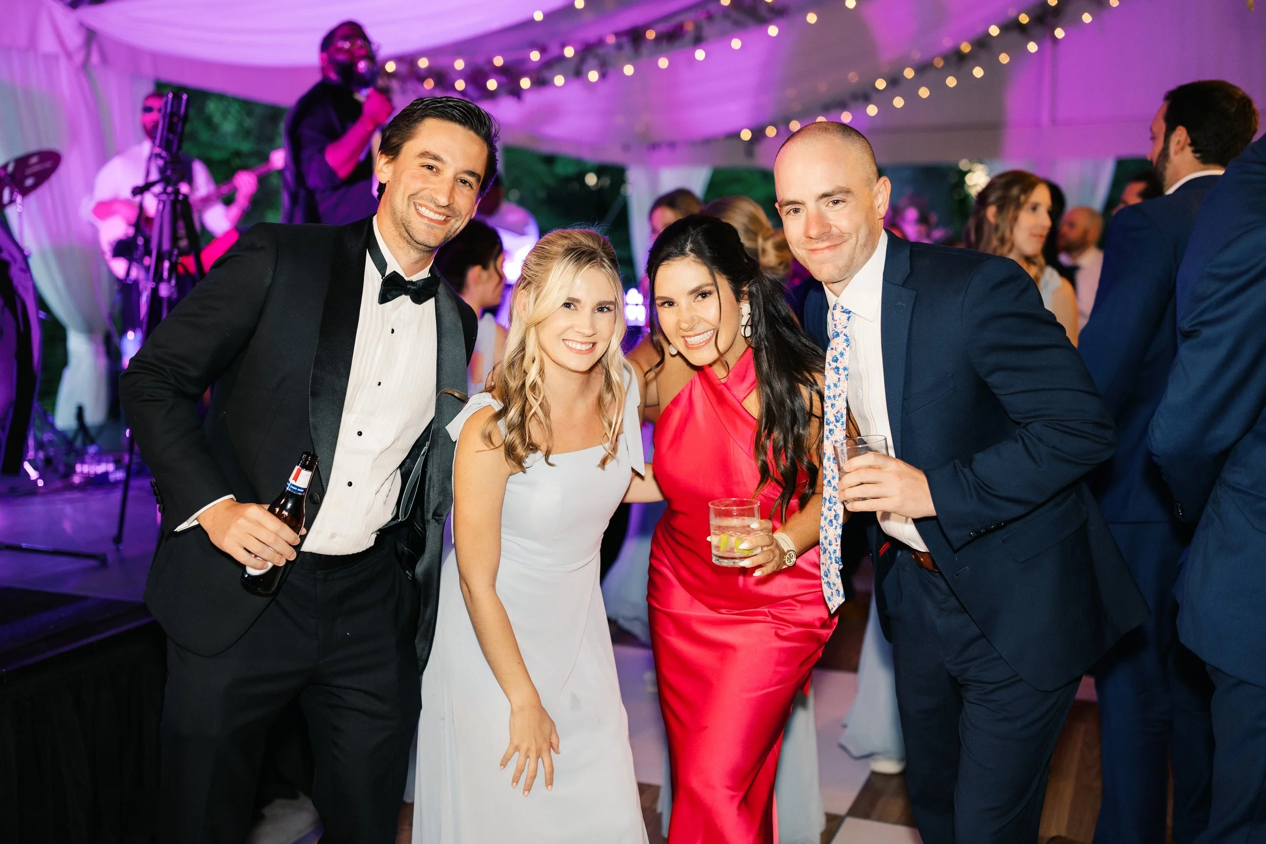 Group of four people dressed in formal attire at a celebration or wedding reception. They are smiling, holding drinks, and standing under string lights with a band performing in the background.