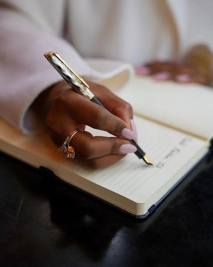 A person writing in a notebook with a gold and black fountain pen, wearing rings, with a blurred background.