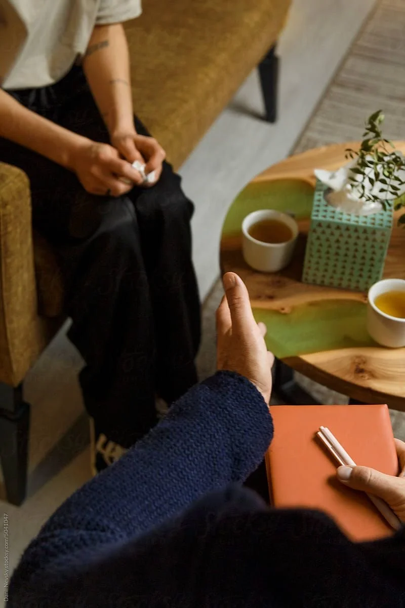 Two people sitting at a small table with tea cups; one person has a notepad and pen, the other is holding a tissue, with a tissue box and a plant on the table.