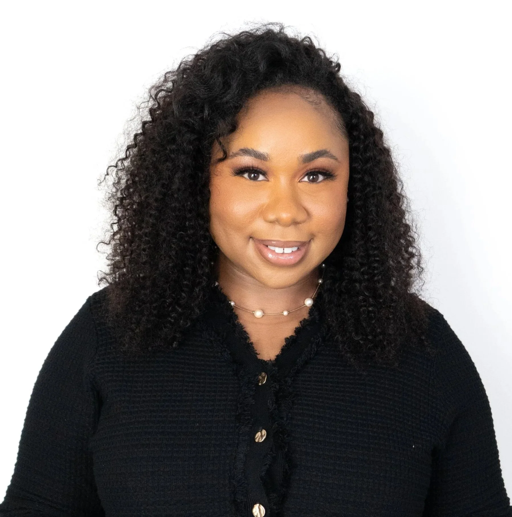 A woman with curly black hair and a warm smile, wearing a black top and a pearl necklace, standing against a white background.