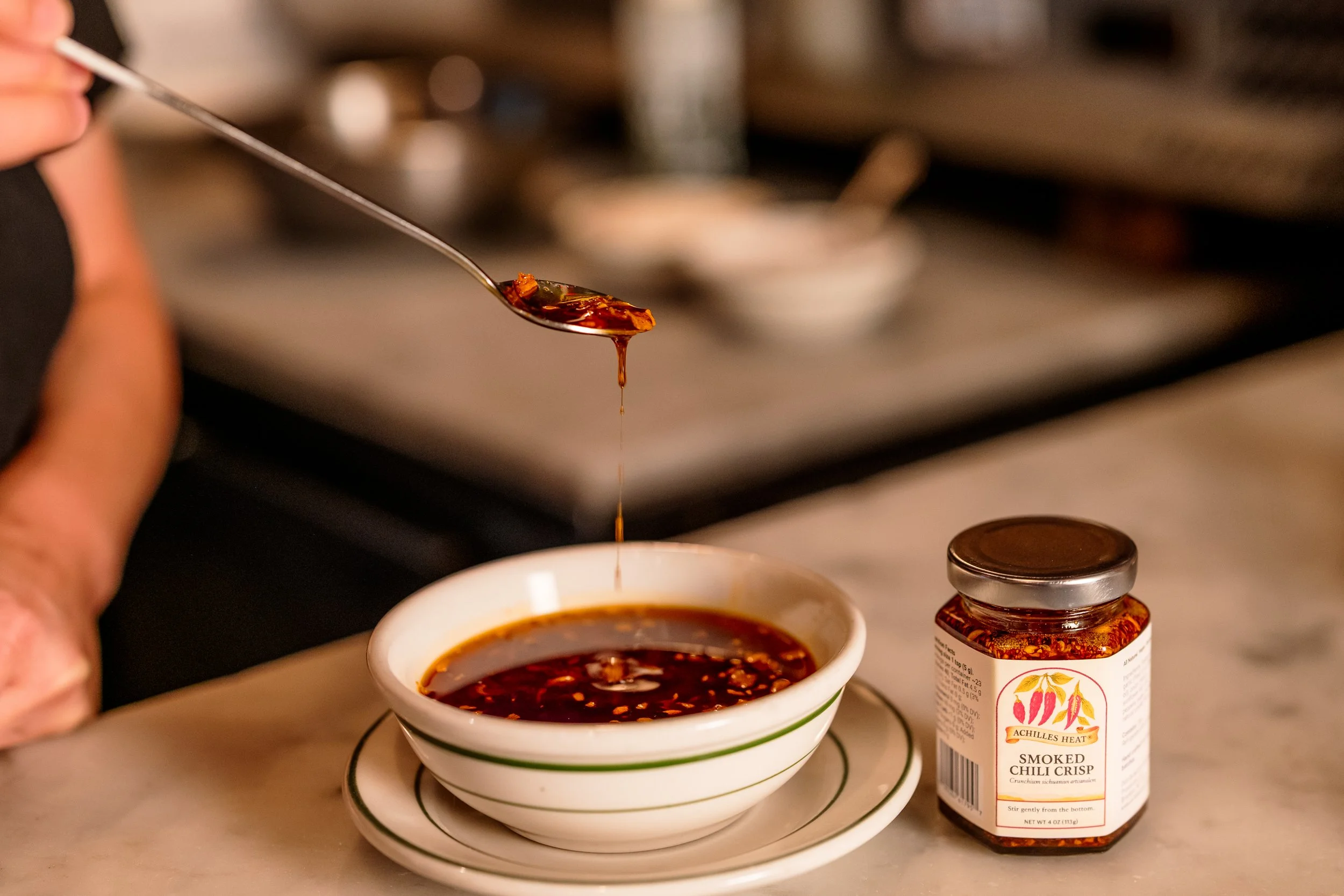 A spoon dripping with hot chili sauce over a bowl of chili sauce on a saucer, with a jar of smoked chili crisp on a marble countertop.