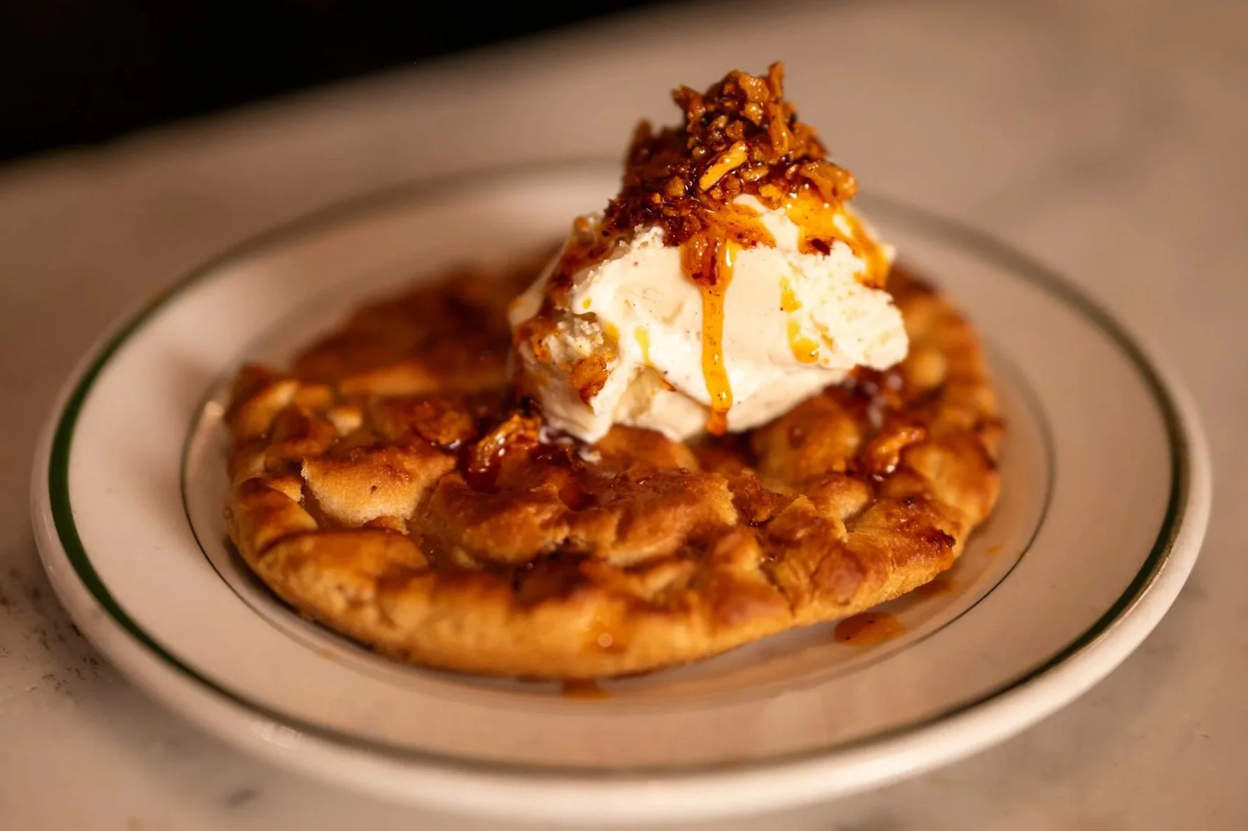 Close-up of apple pie with a scoop of vanilla ice cream, caramel sauce, and chopped nuts on top, served on a white plate.
