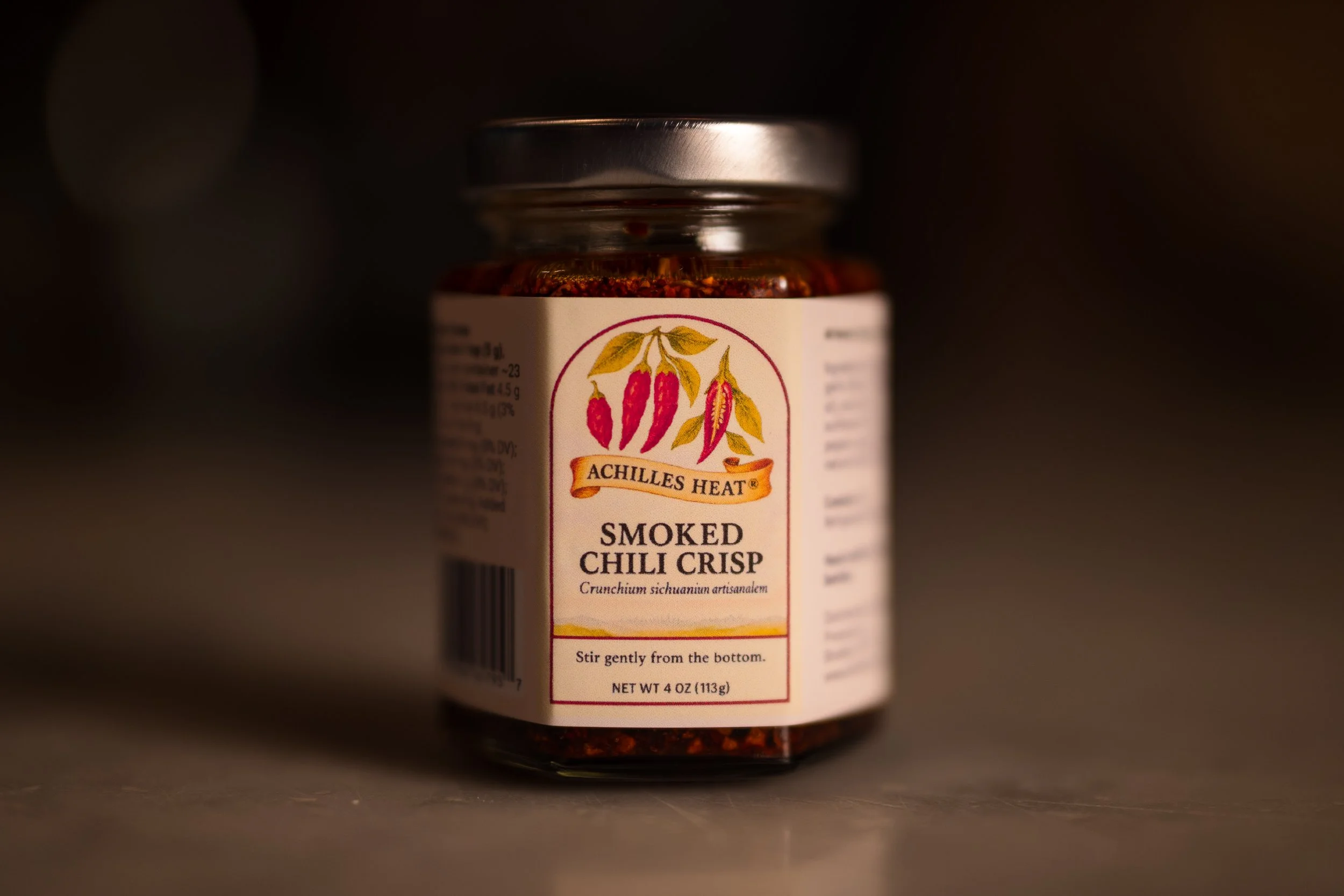 A jar of Achilles Heat Smoked Chili Crisp with a label featuring chili peppers and instructions to stir gently, containing 4 ounces of chili seasoning.