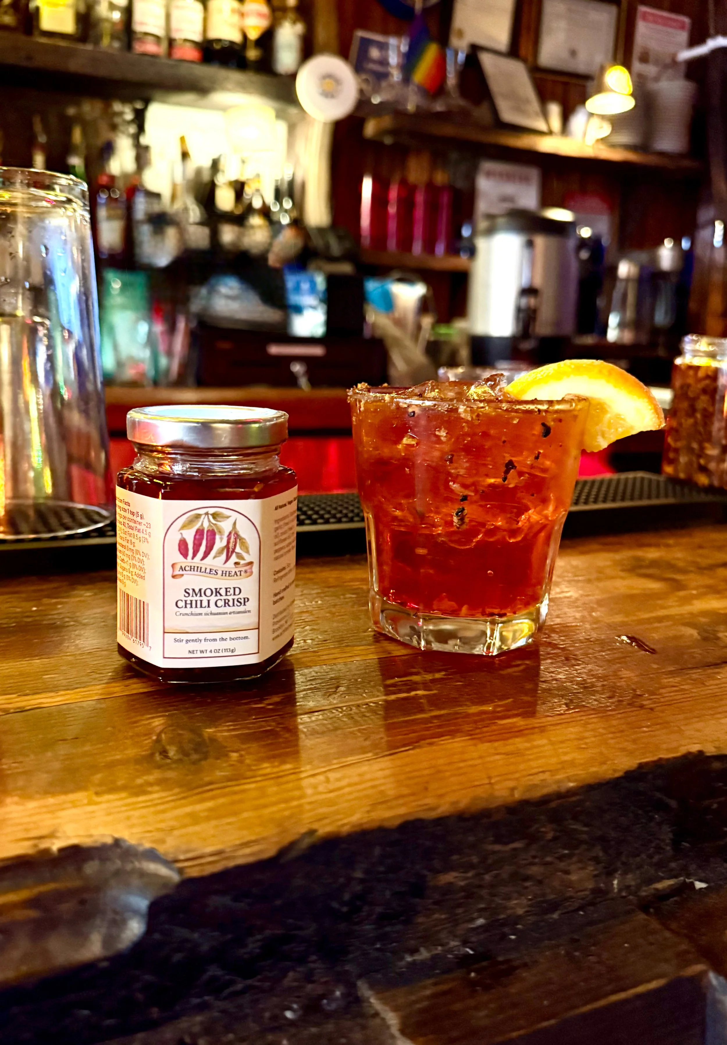 A glass of spicy bloody Mary garnished with a lemon wedge, a jar of smoked chili spice, and a bar counter in the background.