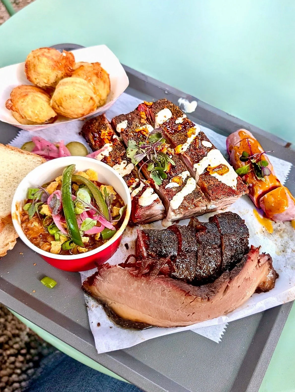 A tray of assorted meats including brisket and sausage, along with fried dough balls and a small bowl of pickled vegetables, served on a metal tray with green background.