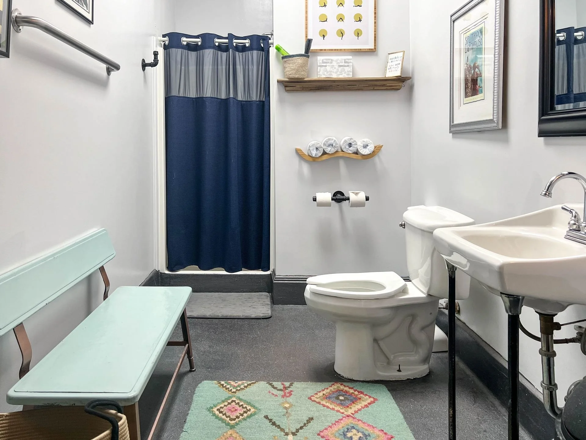Small bathroom with a toilet, sink, and shower with a blue curtain. Light-colored walls decorated with framed artwork, a wall shelf with toilet paper rolls, and a small padded bench. A patterned rug on the gray floor.