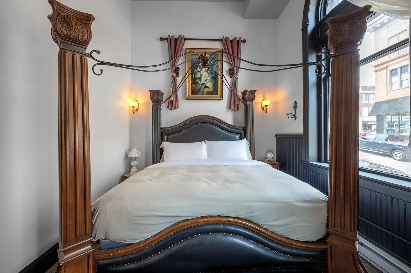 Elegant hotel bedroom with a large four-poster bed, dark wood and leather headboard, ornate wooden posts, pink curtains, artwork above the bed, nightstand with a clock, and large windows showing a street view.