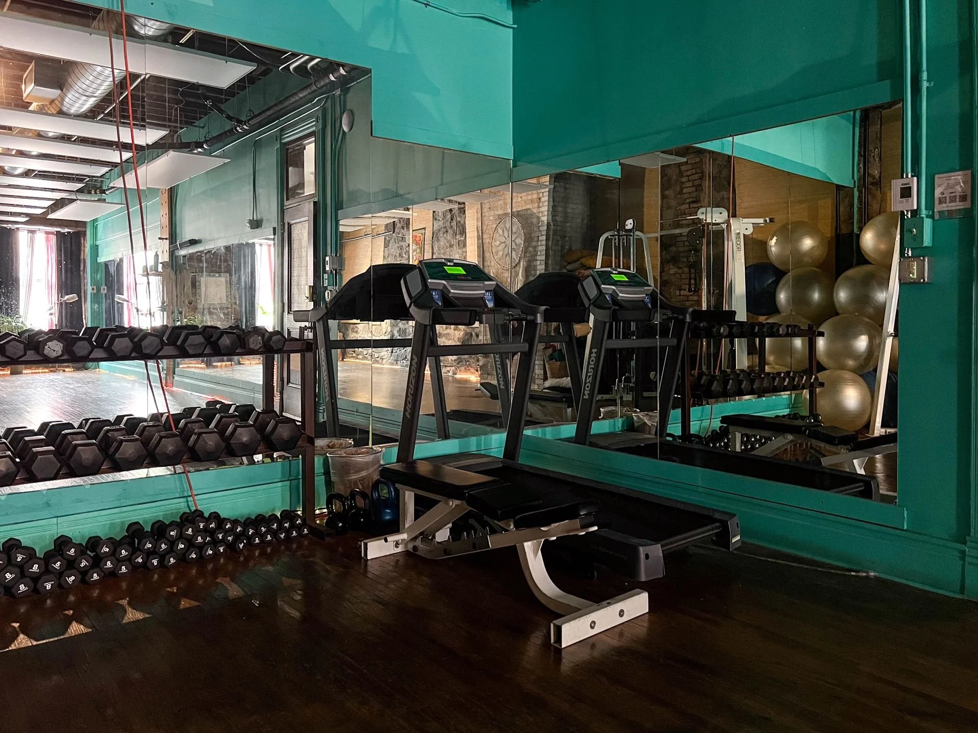 Gym with dumbbells, kettlebells, treadmill, exercise ball, and mirrored wall with exposed brick and industrial ceiling.