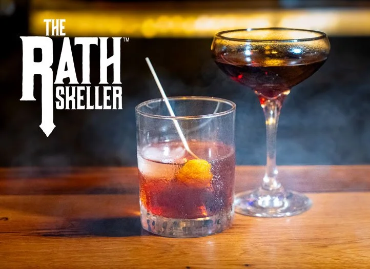 Two cocktails on a wooden bar counter with a dark background, one in a short glass with an orange garnish and a straw, the other in a coupe glass with red wine, and the text "The Bath Skeller" in the top left corner.