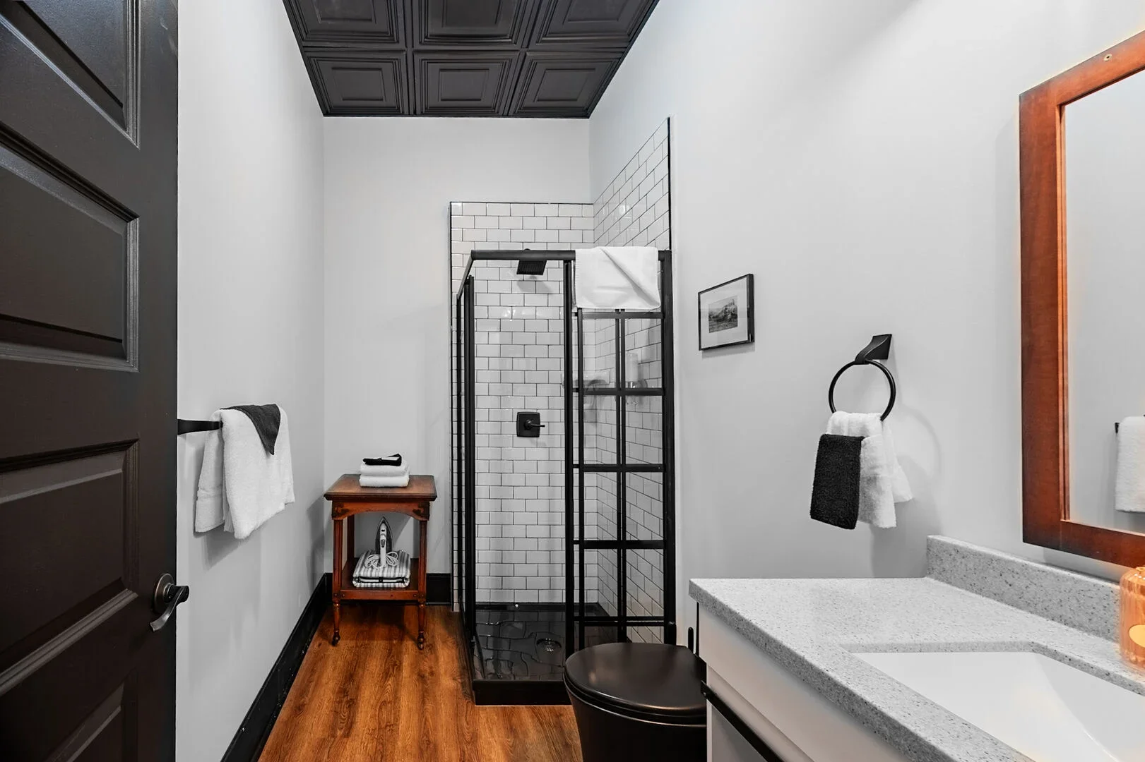 Modern bathroom with black framed glass shower enclosure, white subway tile interior, black fixtures, wooden floor, gray and white vanity with granite countertop, black and white towels, black wall-mounted towel rings, small wooden side table with towels, framed artwork on wall, dark door, and ceiling with black paneling.
