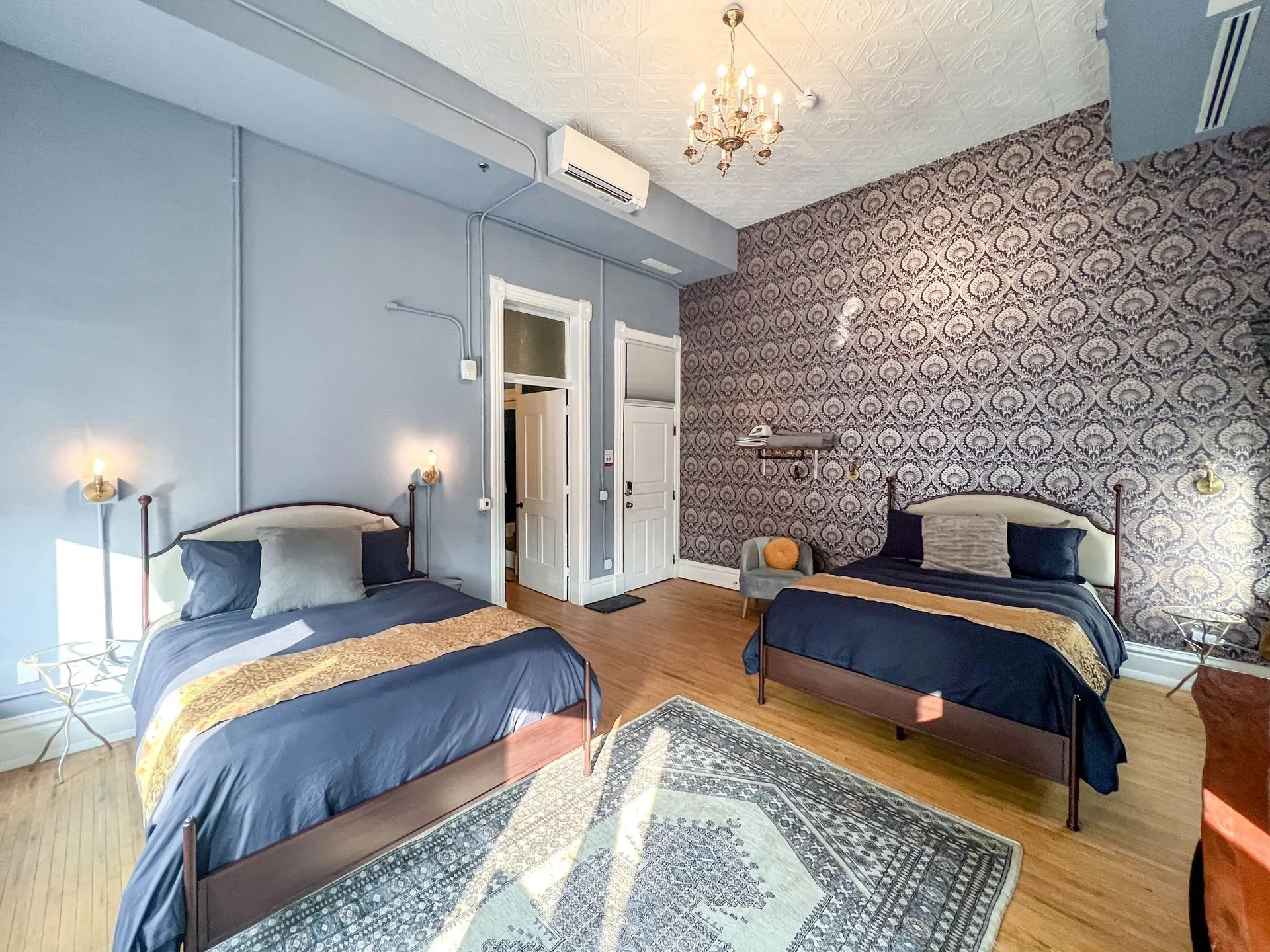 A vintage-style hotel room with two twin beds, each with dark bedspreads and pillows, a patterned accent wall, a chandelier hanging from the ceiling, a small gray armchair with an orange pillow, a wooden nightstand, and a patterned rug on a wooden floor.