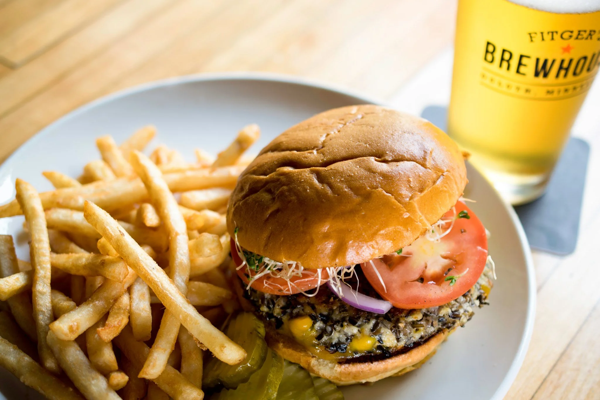 A plate with French fries, a cheeseburger with tomato, onion, and sprouts, and pickles, with a glass of beer in the background.