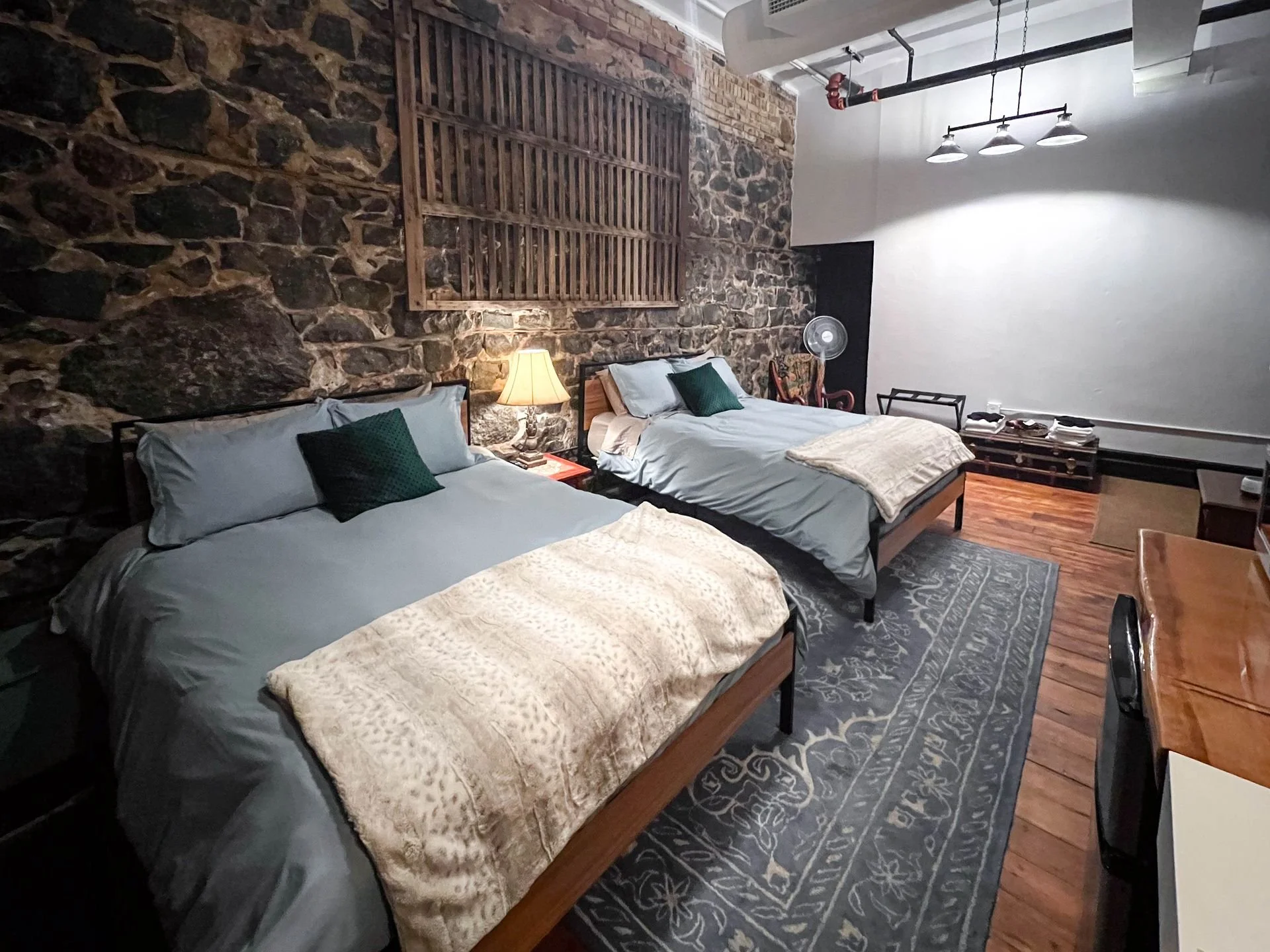 Two beds with gray bedding and green pillows in a rustic hotel room with stone wall and wooden accents.