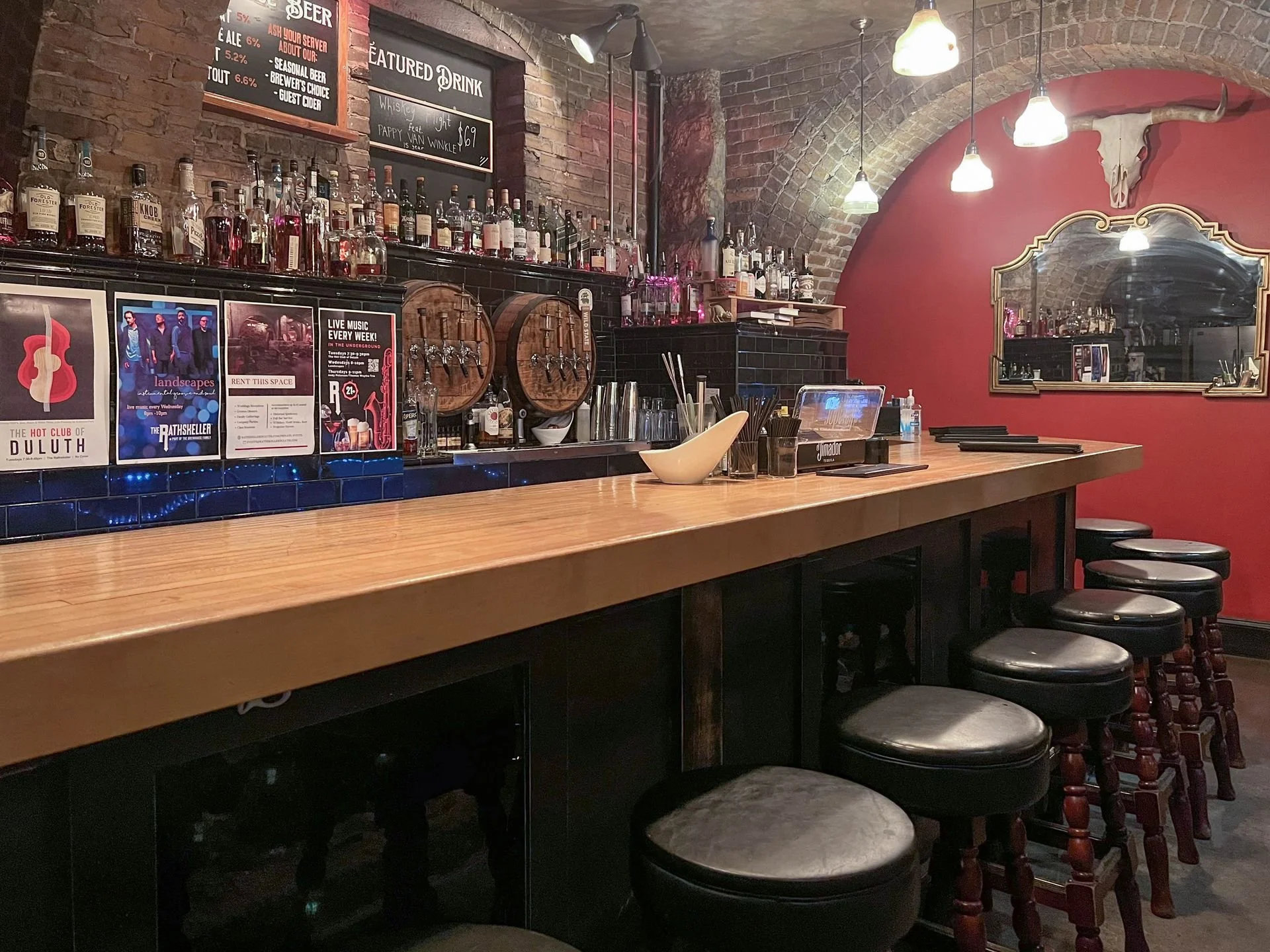 Bar with a wooden counter, black stools with cushioned seats, and a backdrop of liquor bottles on shelves, a mirror, and a burgundy wall with a skull and cow skull decor.