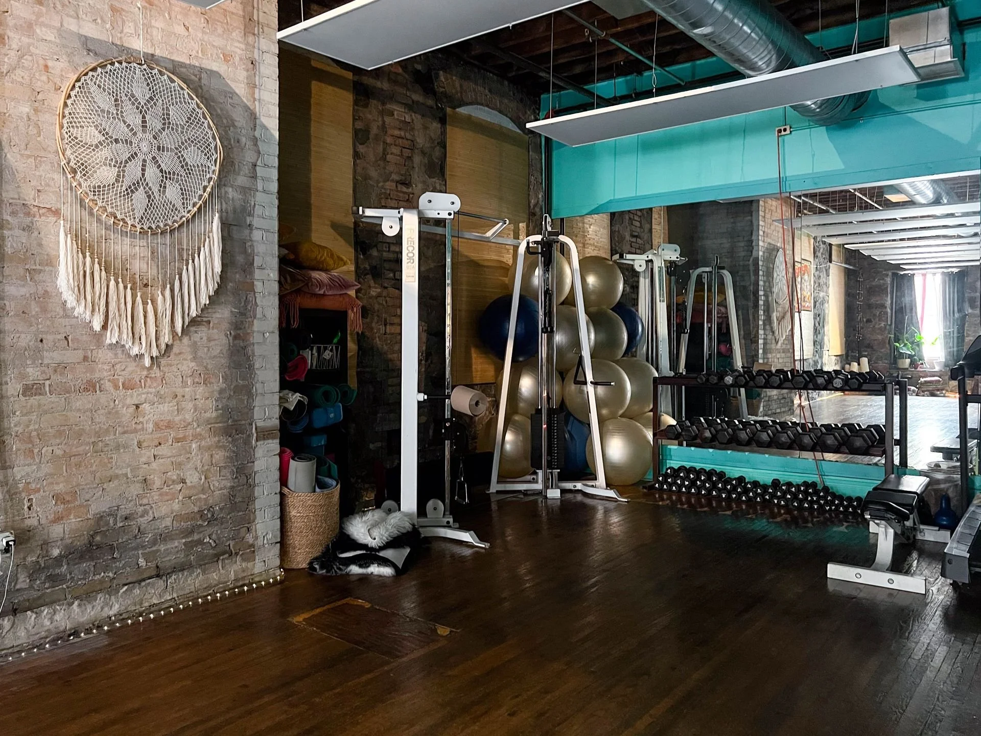 Interior of a gym with brick walls and wooden floors, featuring exercise equipment such as balance balls, dumbbells, and resistance bands, and decorative wall hanging.