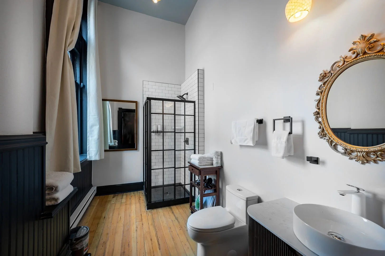 A bathroom with white walls, dark wood paneling, and a wooden floor. Features include a glass shower with black framing and white subway tiles, a toilet, a white sink on a dark cabinet, a large ornate golden framed mirror, a small wooden shelving unit with towels and toiletries, towel racks with white towels, a window with beige curtains, a wall-mounted light fixture, and a small mirror on the wall.