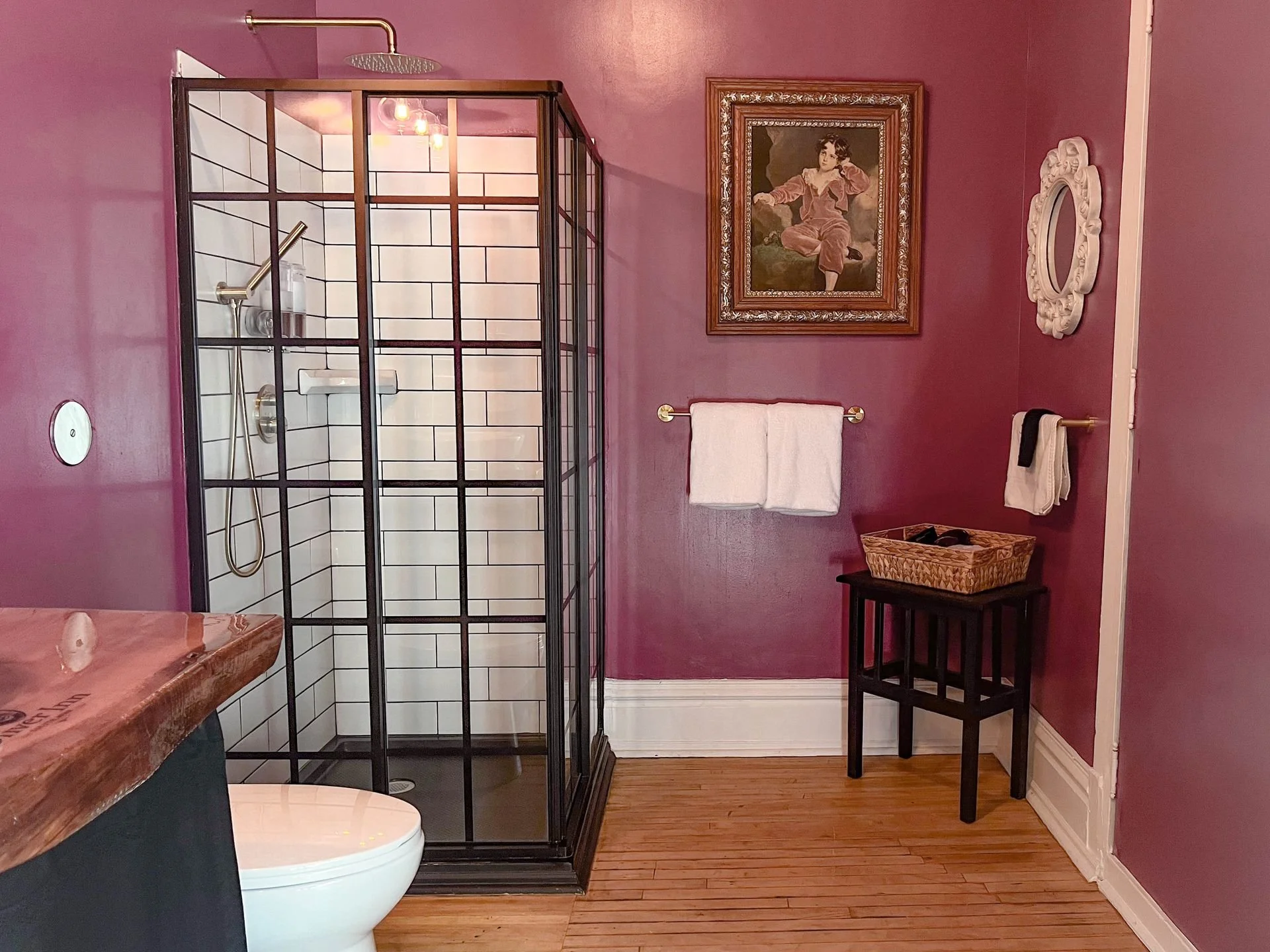 A bathroom with purple walls, a glass shower with black framing, a white toilet, a wooden countertop, framed artwork, a mirror, a towel rack with towels, a small black table, and a basket.