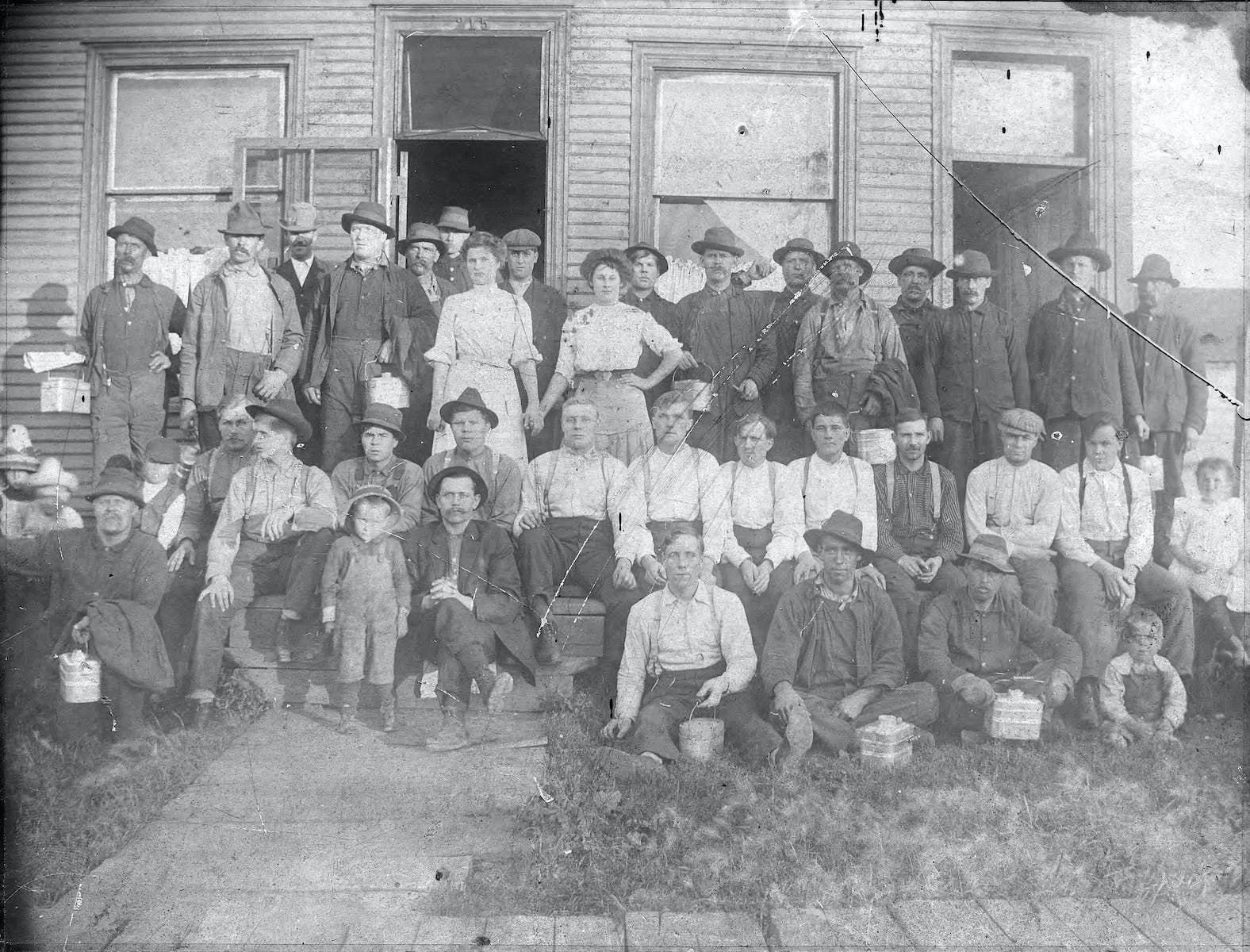 Black and white photograph of a large group of people, men, women, and children, posing in front of a wooden house. The group is arranged in rows, some seated and some standing, with various expressions. Many are dressed in early 20th-century clothing, including hats and jackets.