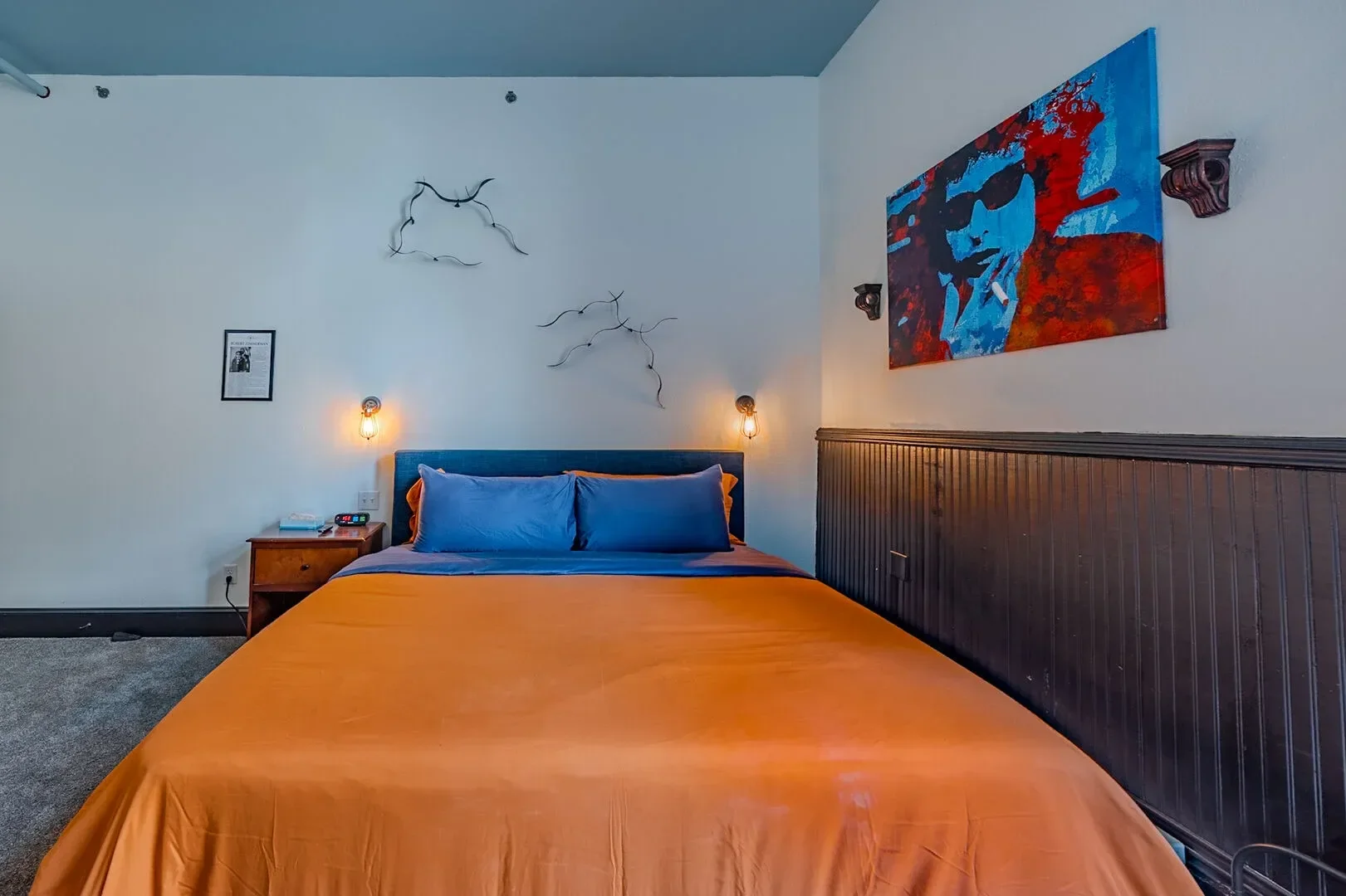 A bedroom with a bed that has an orange bedspread and blue pillows, a wooden nightstand with a telephone, and wall-mounted lights. The wall features a colorful abstract portrait and decorative metal branches.