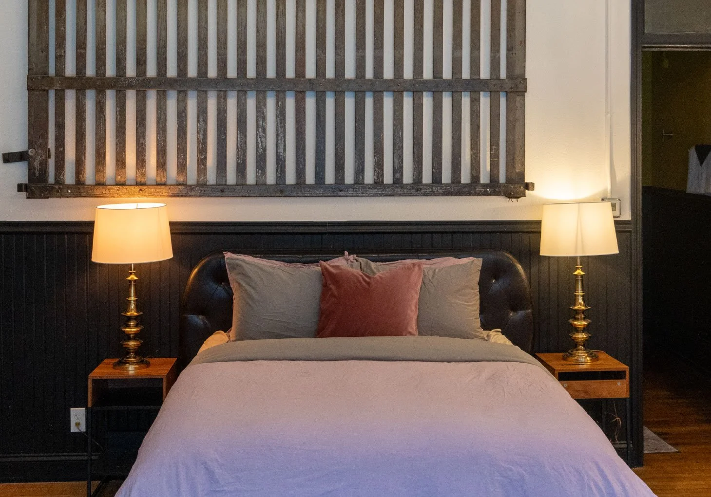 A bedroom with a bed, pillows, and two lamps on nightstands on either side. There is a wooden slatted headboard above the bed, and a doorway to the right.