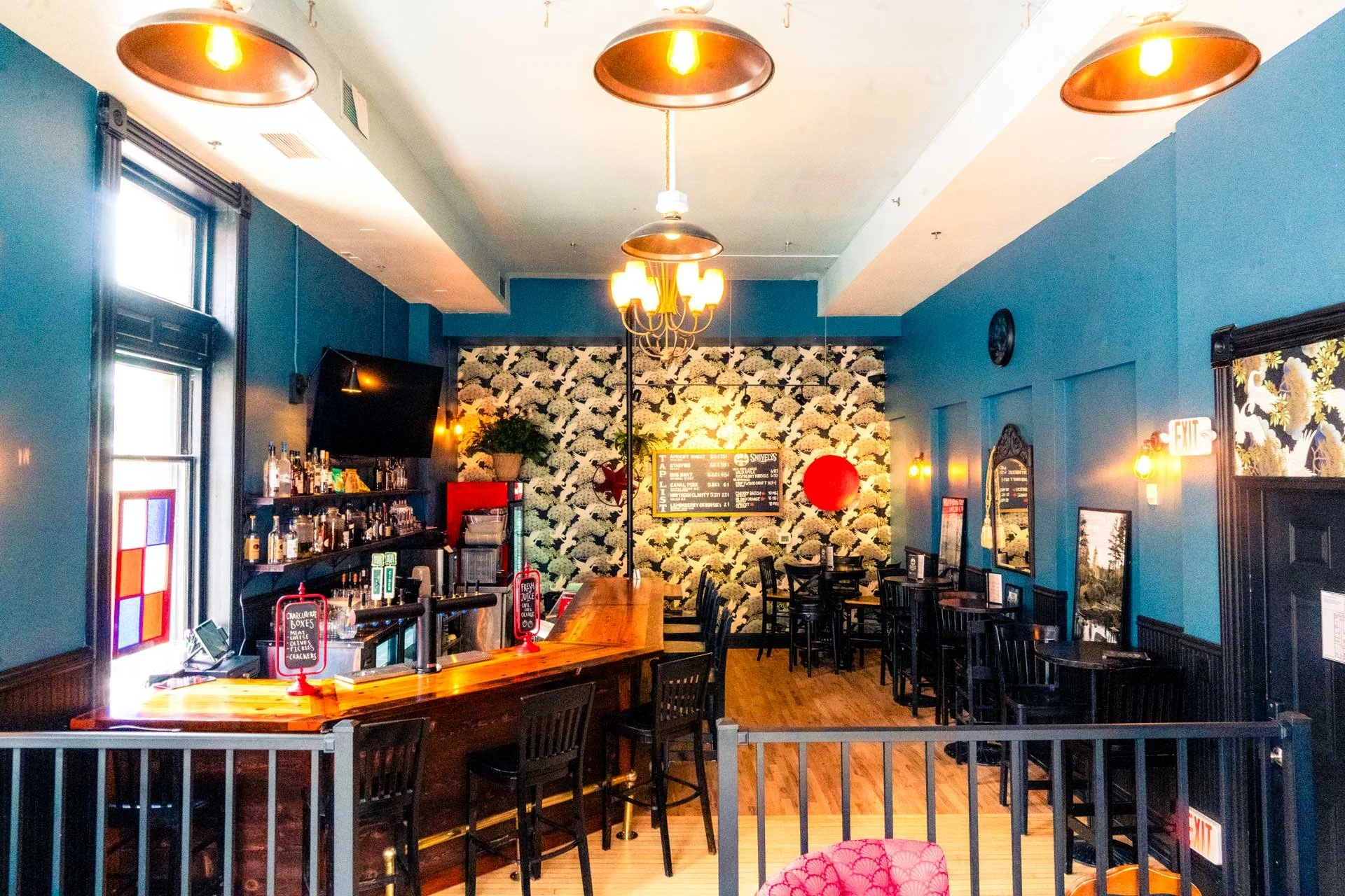 Interior of a cozy bar with turquoise walls and a mix of high and low tables, a decorated back wall with floral wallpaper, and warm lighting fixtures.