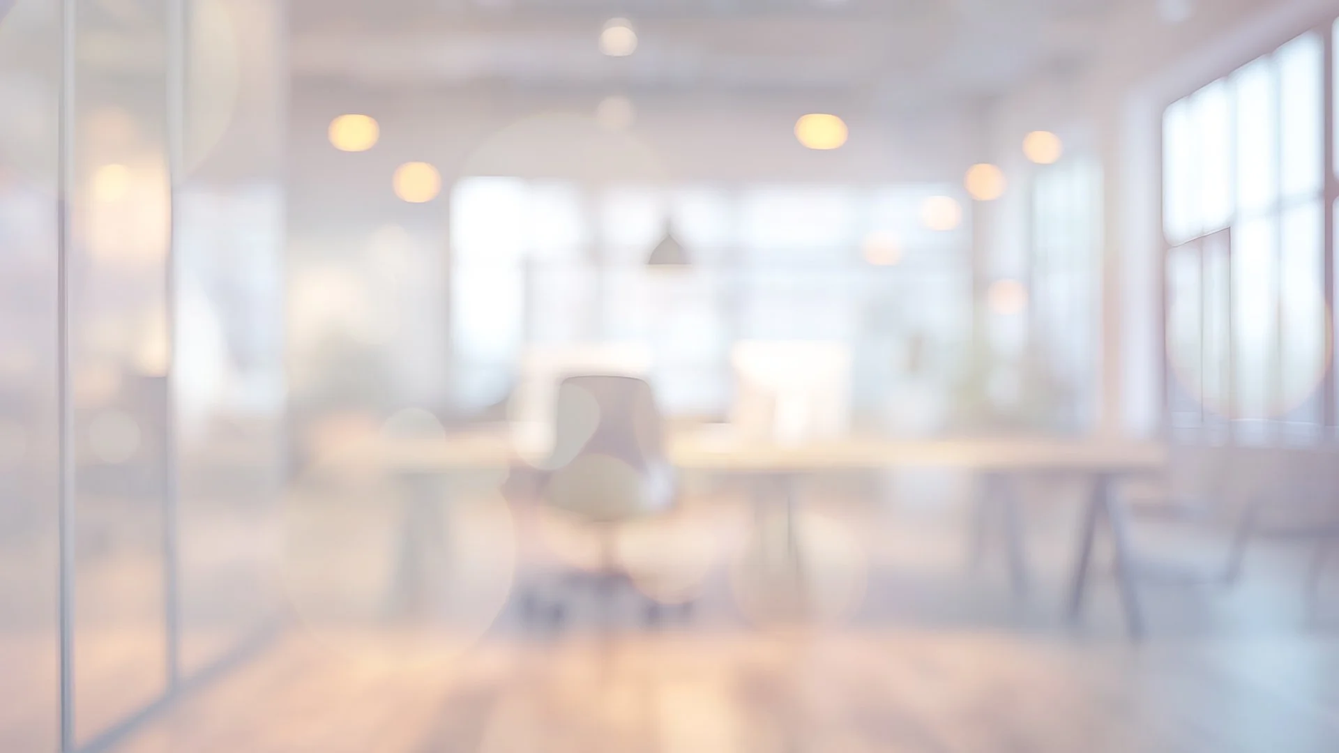 Blurred view of an office space seen through a glass window, with hanging lights and large windows allowing natural light to fill the room.