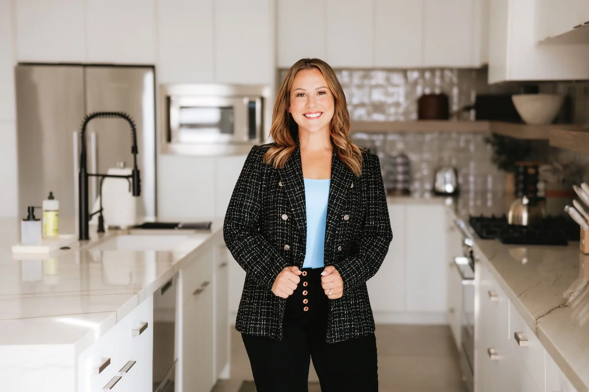 A woman with light brown hair smiling and standing in a modern kitchen, wearing a black checkered blazer and black pants.
