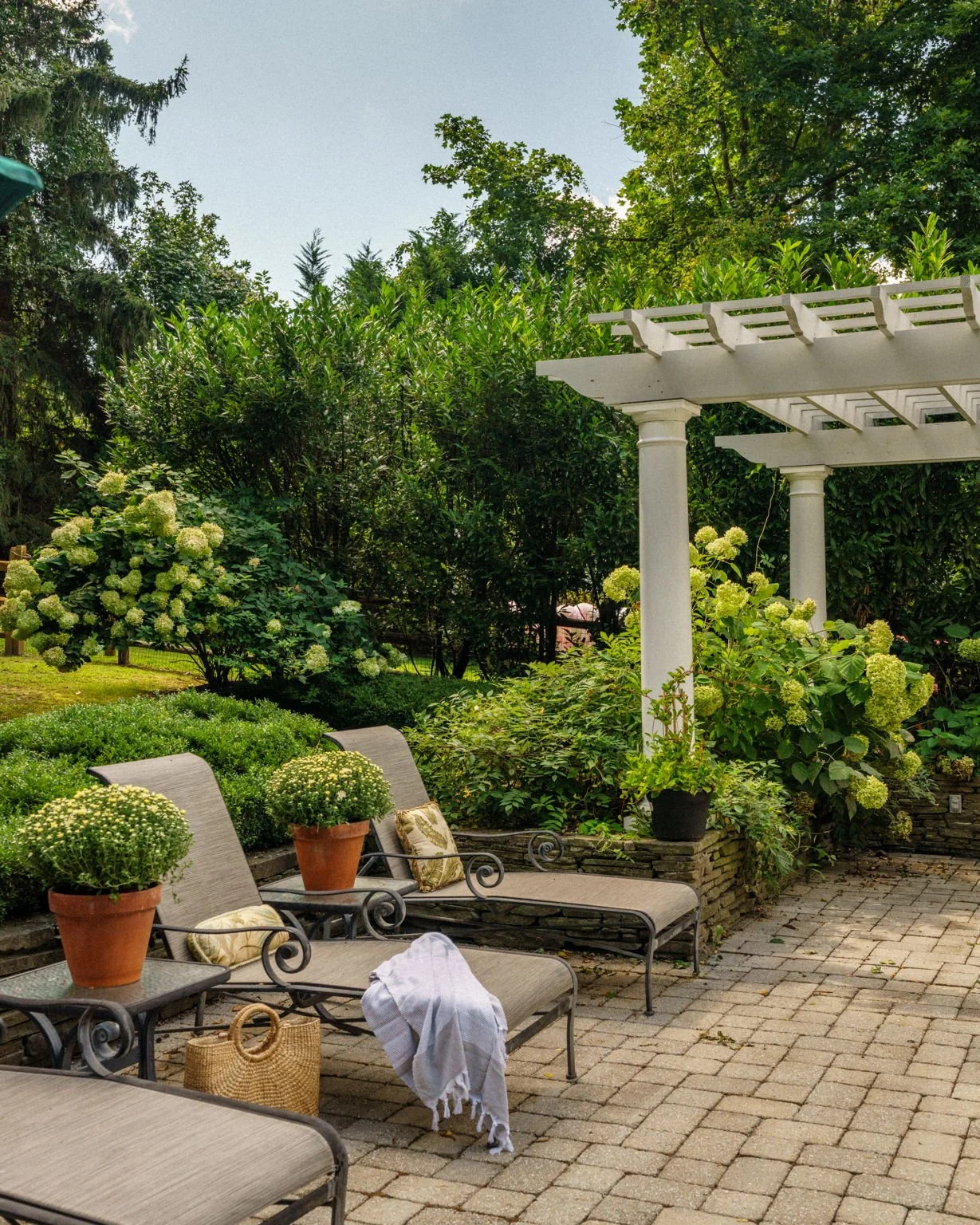 A backyard patio with three lounge chairs, potted plants, and a white pergola surrounded by greenery and flowering bushes.