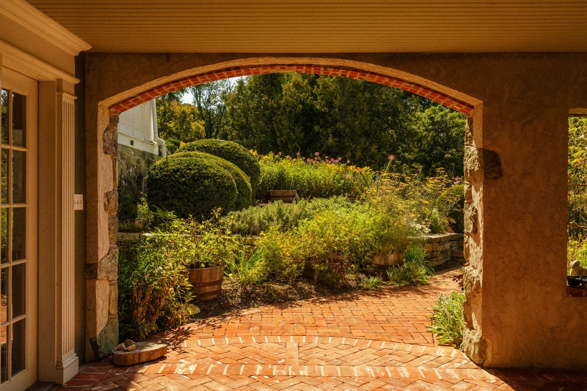 View from a covered porch looking onto a garden with lush green bushes, plants, and trees, with a brick-paved walkway and a stone wall in the background.