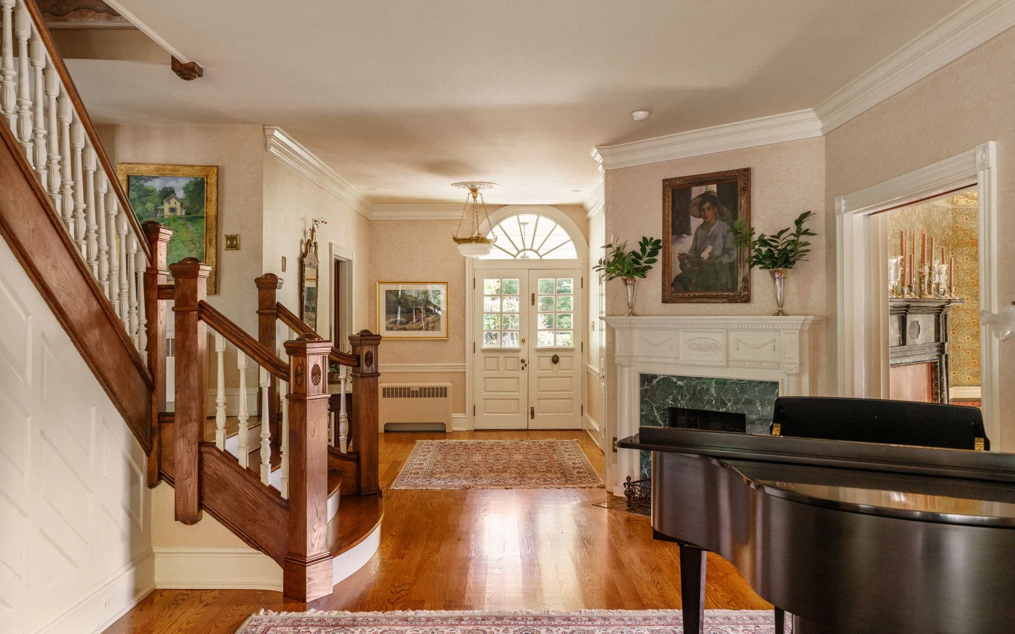 Interior view of a spacious home entrance with hardwood floors, a grand piano on the right, a fireplace with a portrait above, and a front door with glass panels and a semi-circle window above.