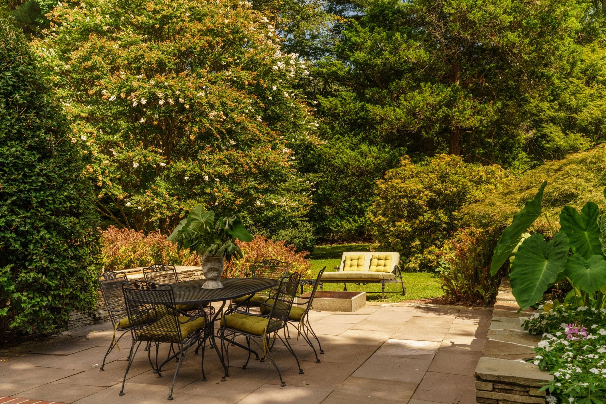 A garden patio with outdoor furniture, including a black metal table with a large vase of flowers, six chairs with green cushions, and a yellow cushioned bench, surrounded by lush green trees and plants.