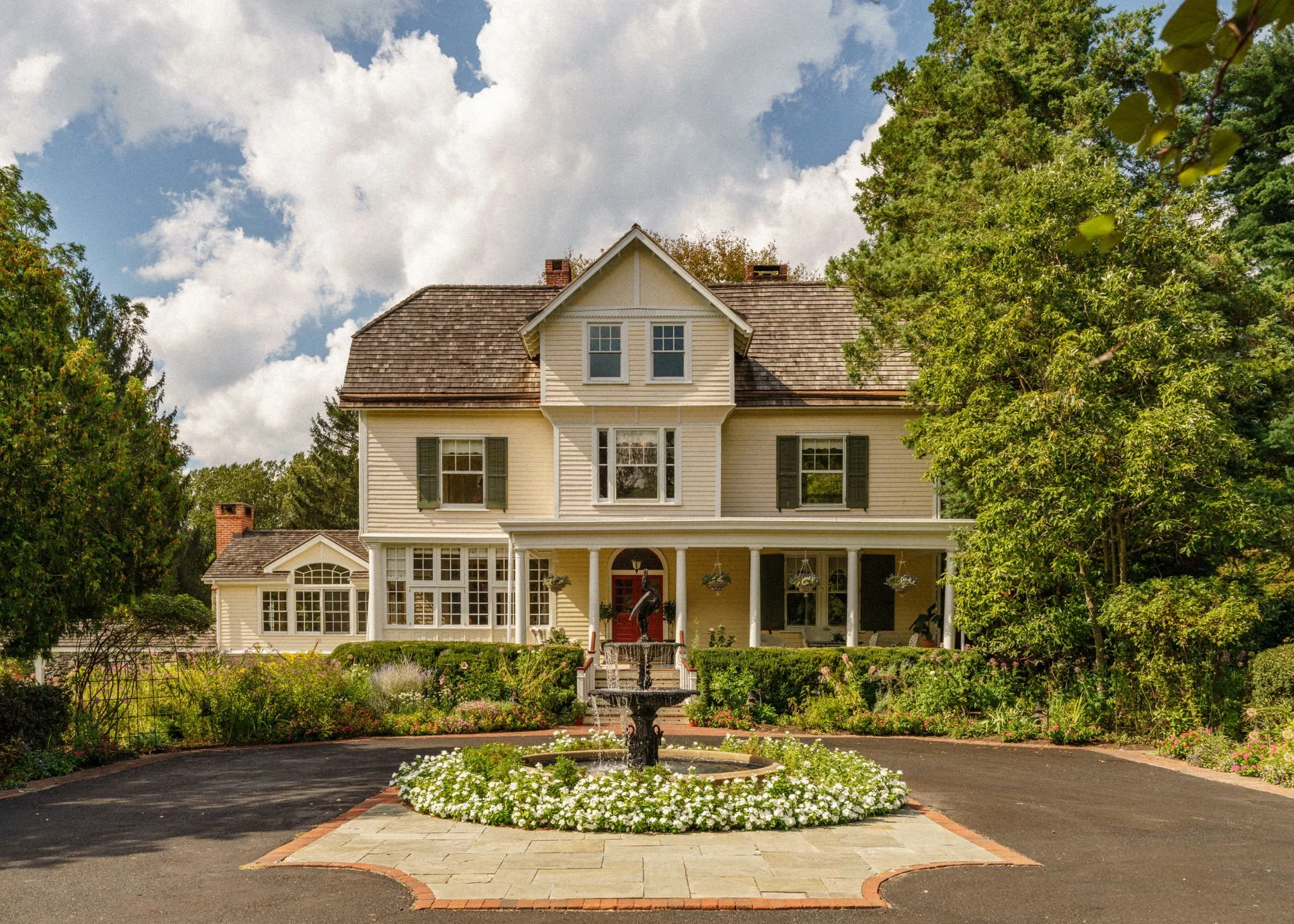 Large, historic house with a front porch, surrounded by a lush garden with a circular driveway, fountain, and trees.