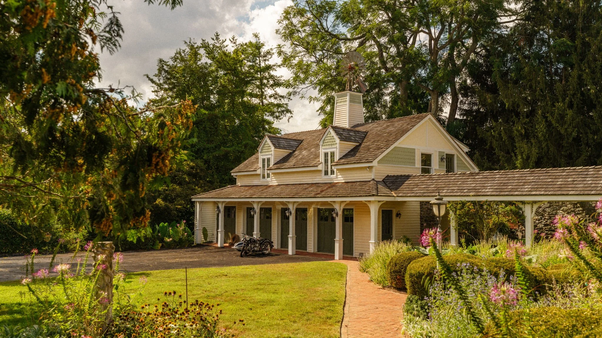 A two-story house with a porch and garage, surrounded by a garden with various plants and flowers, and tall trees in the background.