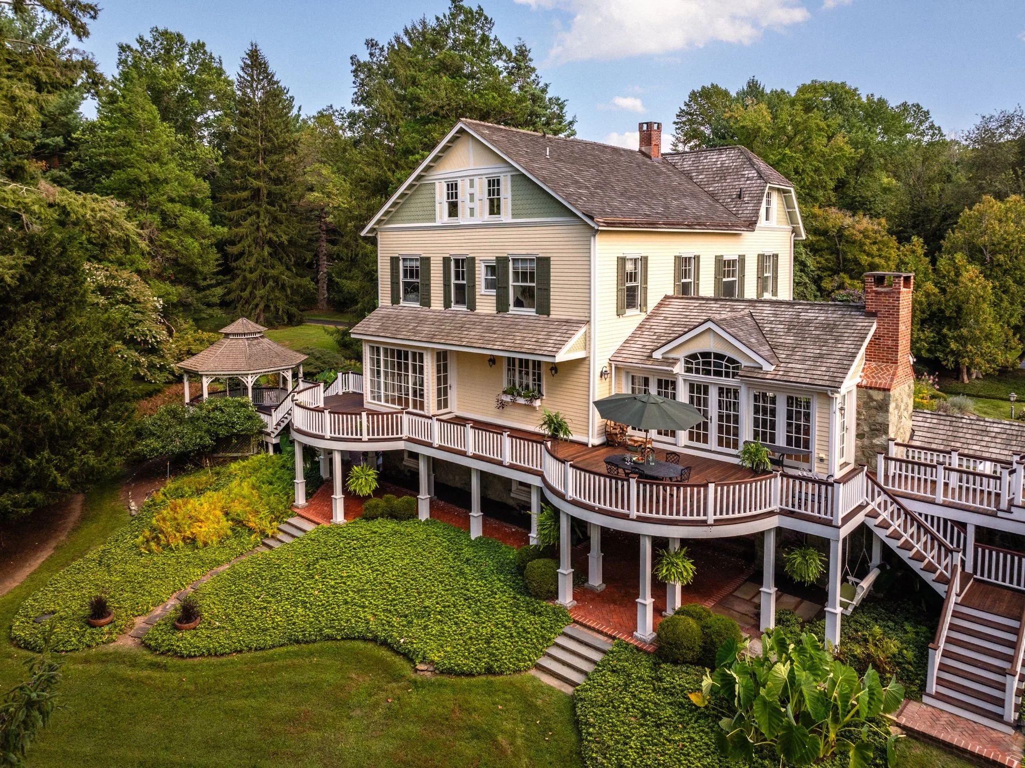 A large, beige, multi-story house with a wrap-around deck and green shutters, surrounded by lush greenery and trees.