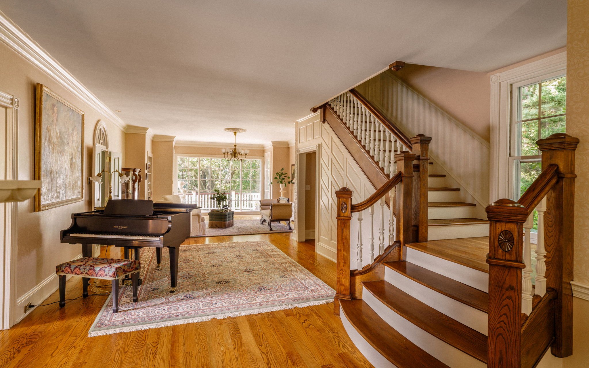 Living room with a grand staircase, a piano with a patterned stool, a large area rug, and a sitting area near large windows overlooking greenery.