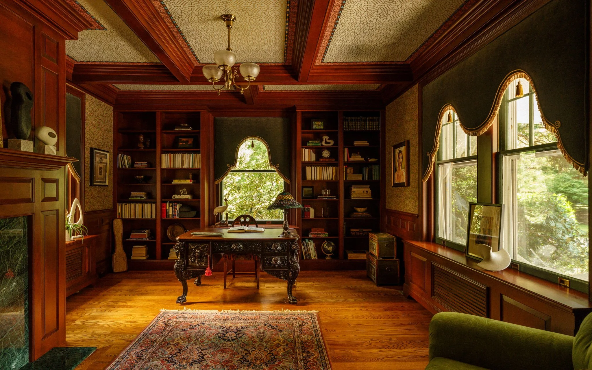 Elegant home library with rich wooden paneling, large windows with ornate valances, a vintage desk with a Tiffany-style lamp, surrounded by bookshelves filled with books and decorative items, and a patterned area rug on hardwood floors.