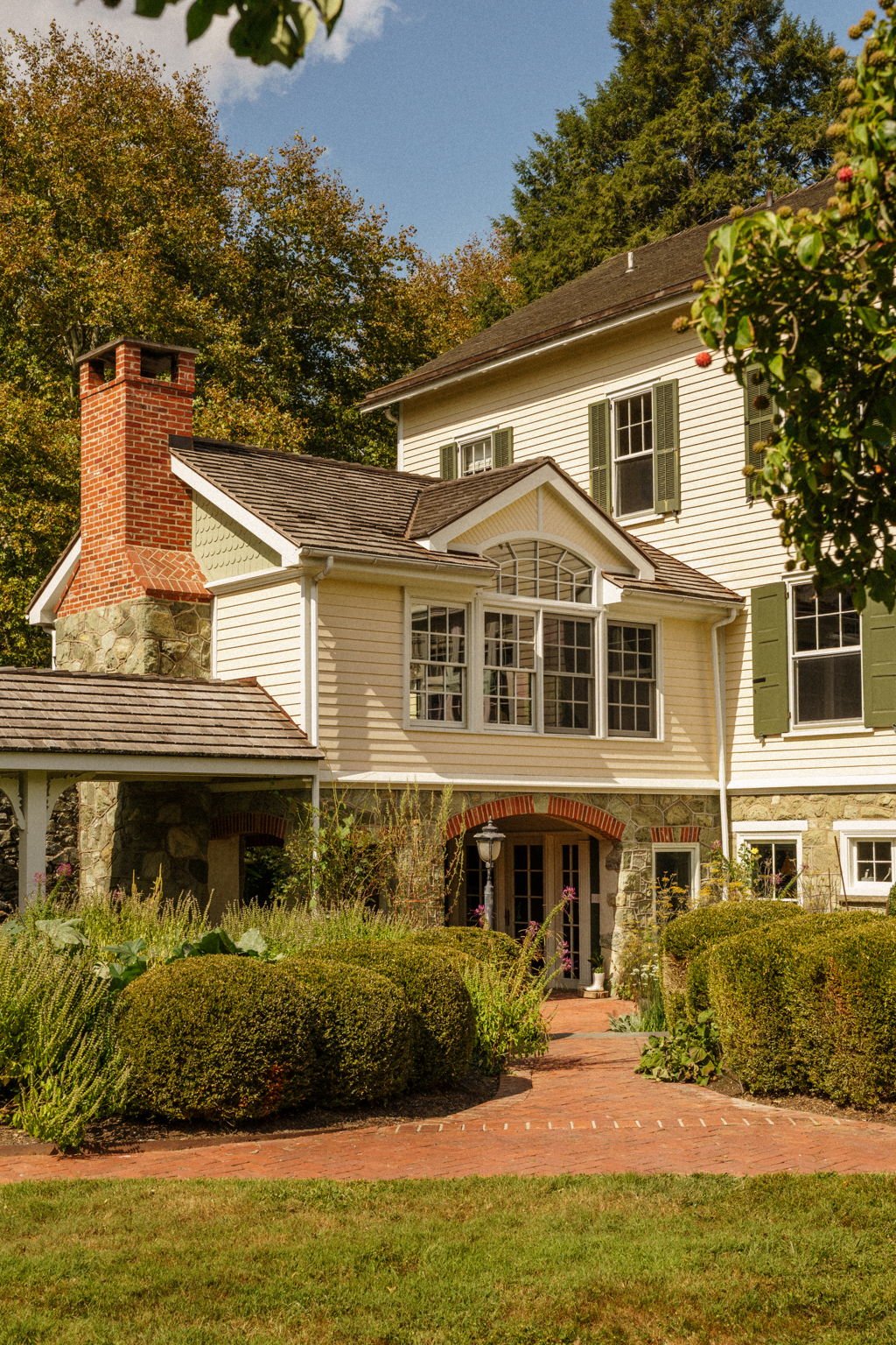 Front view of a two-story house with a stone foundation, white siding, green shutters, and a brick chimney, surrounded by greenery and a brick walkway.