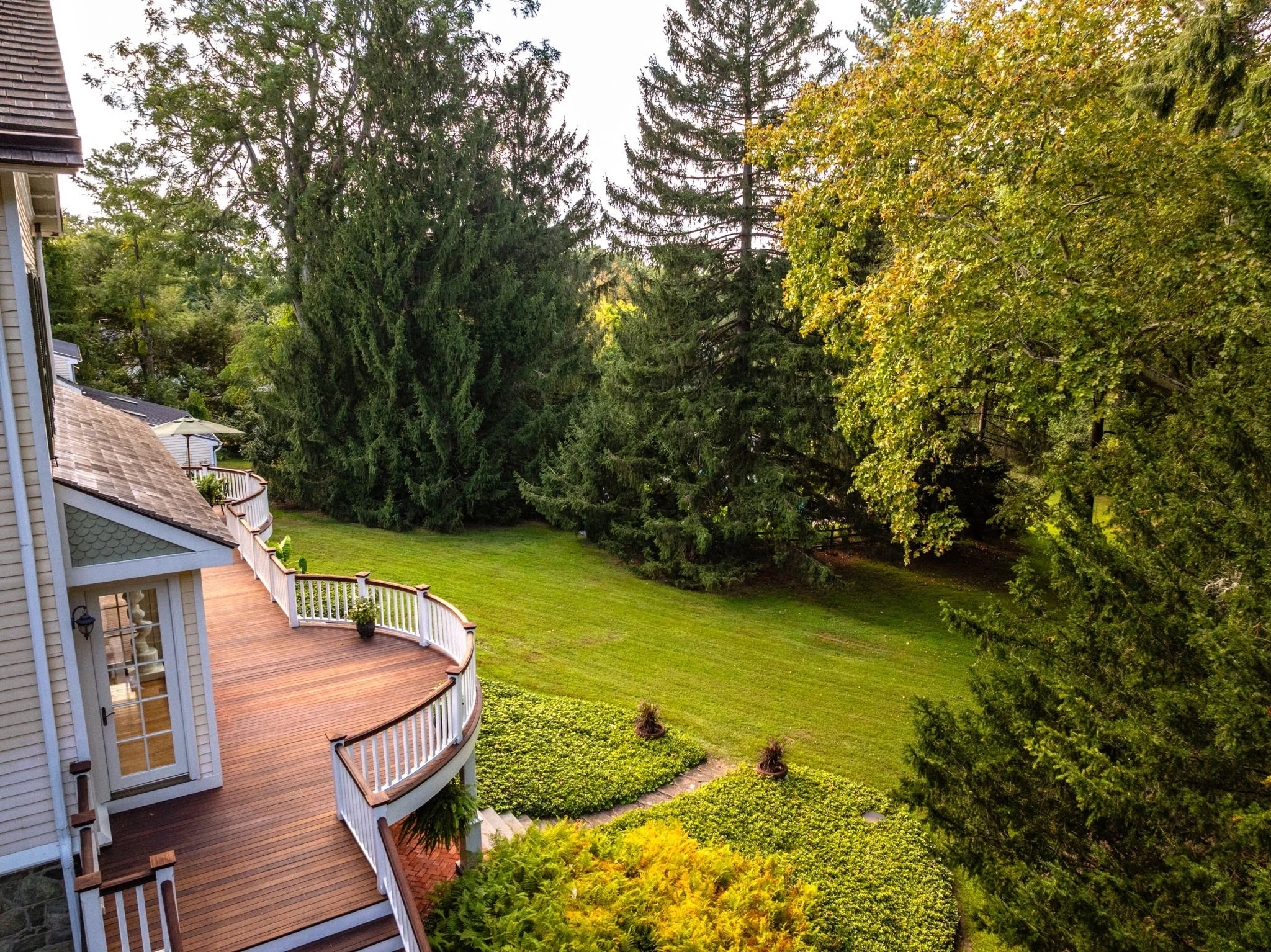 View of a backyard with a wooden deck, lush green lawn, and tall trees surrounding the area.