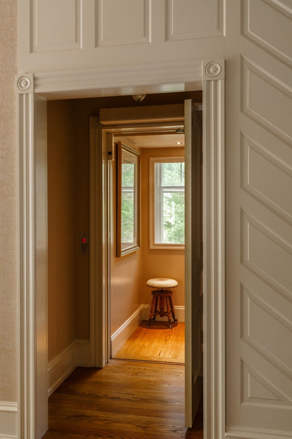View through an open elevator door into a small room with hardwood floors, a window, and a stool.