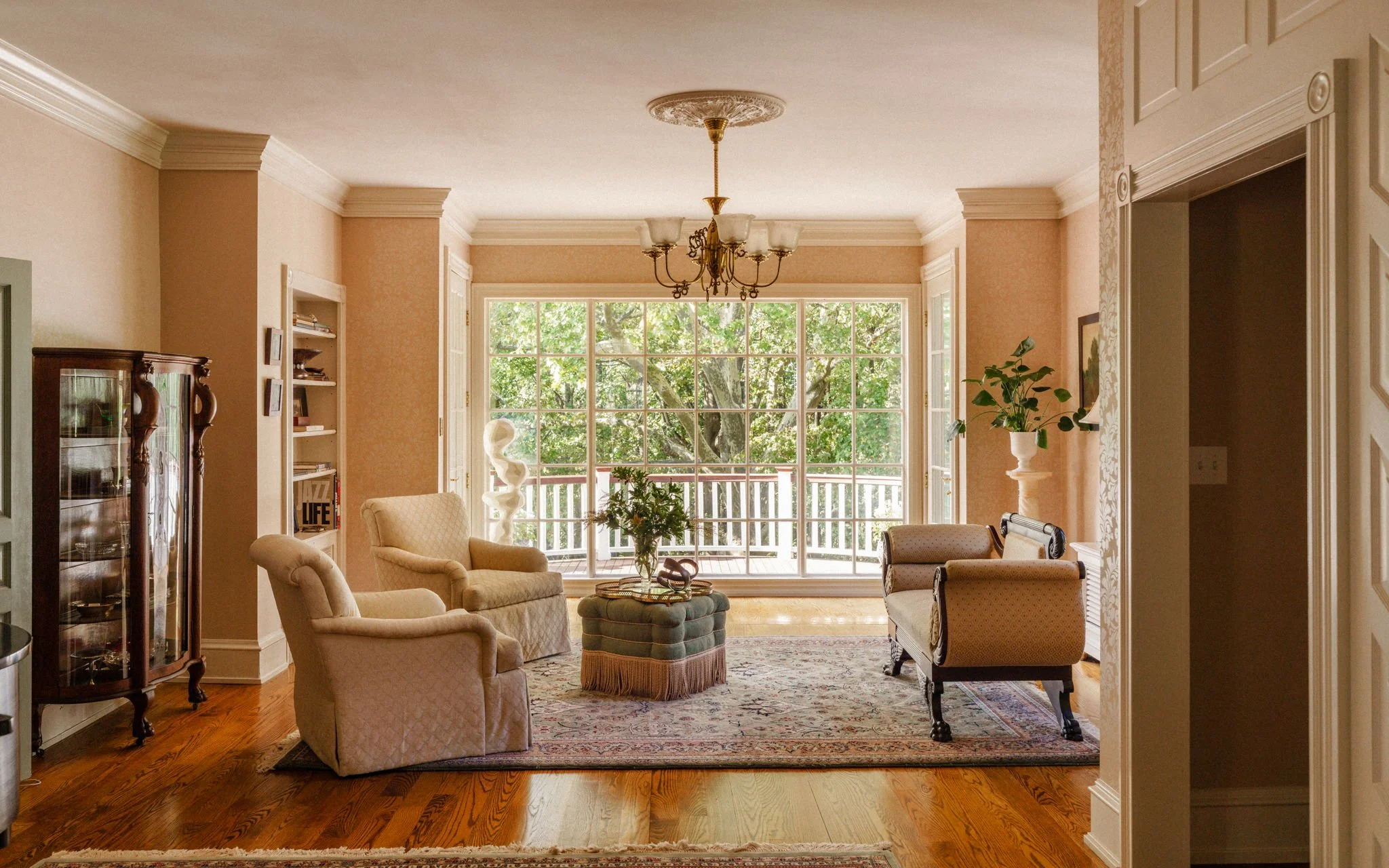 Bright living room with large window overlooking trees, beige and cream furniture, chandelier, and hardwood floors.