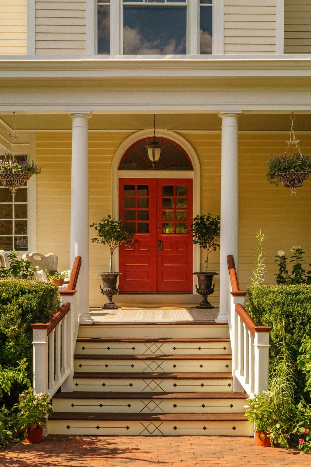 Front porch of a yellow house with red double doors, white columns, hanging flower baskets, potted plants, and a brick walkway.