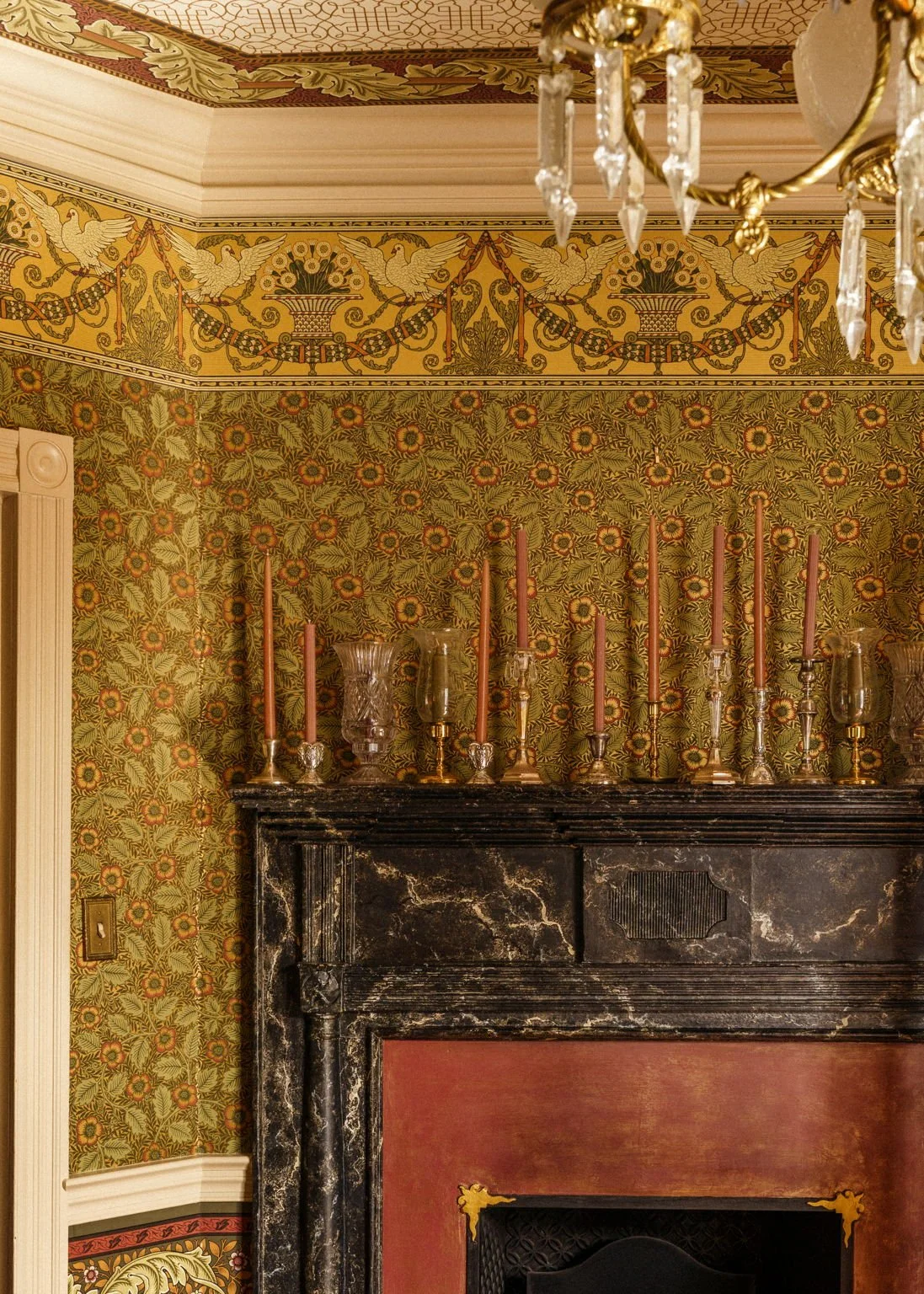 Close-up of a vintage room corner with a black marble fireplace, ornate wallpaper with floral and bird patterns, a decorative chandelier, and a mantel decorated with candles and glass vases.