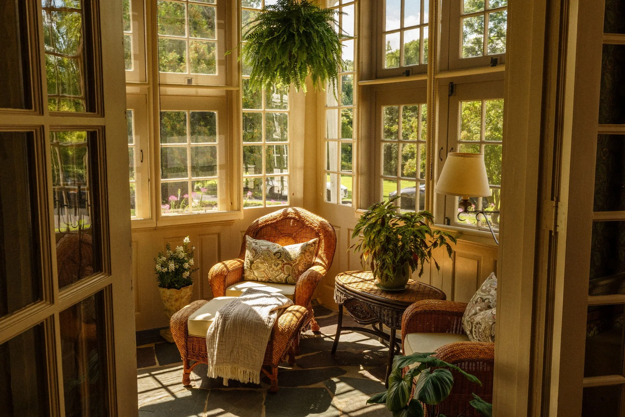 Sunlit cozy sitting area with wicker armchair, floral pillow, throw blanket, side table with potted plant, and floor lamp, surrounded by large windows with greenery outside.