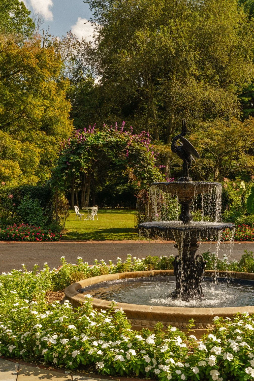 A decorative fountain with water cascading from a bird sculpture on top, surrounded by flowering plants, in a garden setting with trees, a grassy area, and white garden furniture in the background.