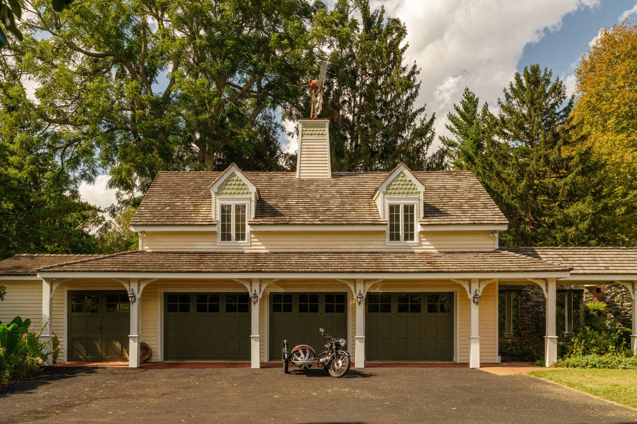 A house with a three-car garage, yellow siding, and a shingled roof is surrounded by trees. There is a bicycle with a sidecar parked in front of the garage, and the sky is partly cloudy.