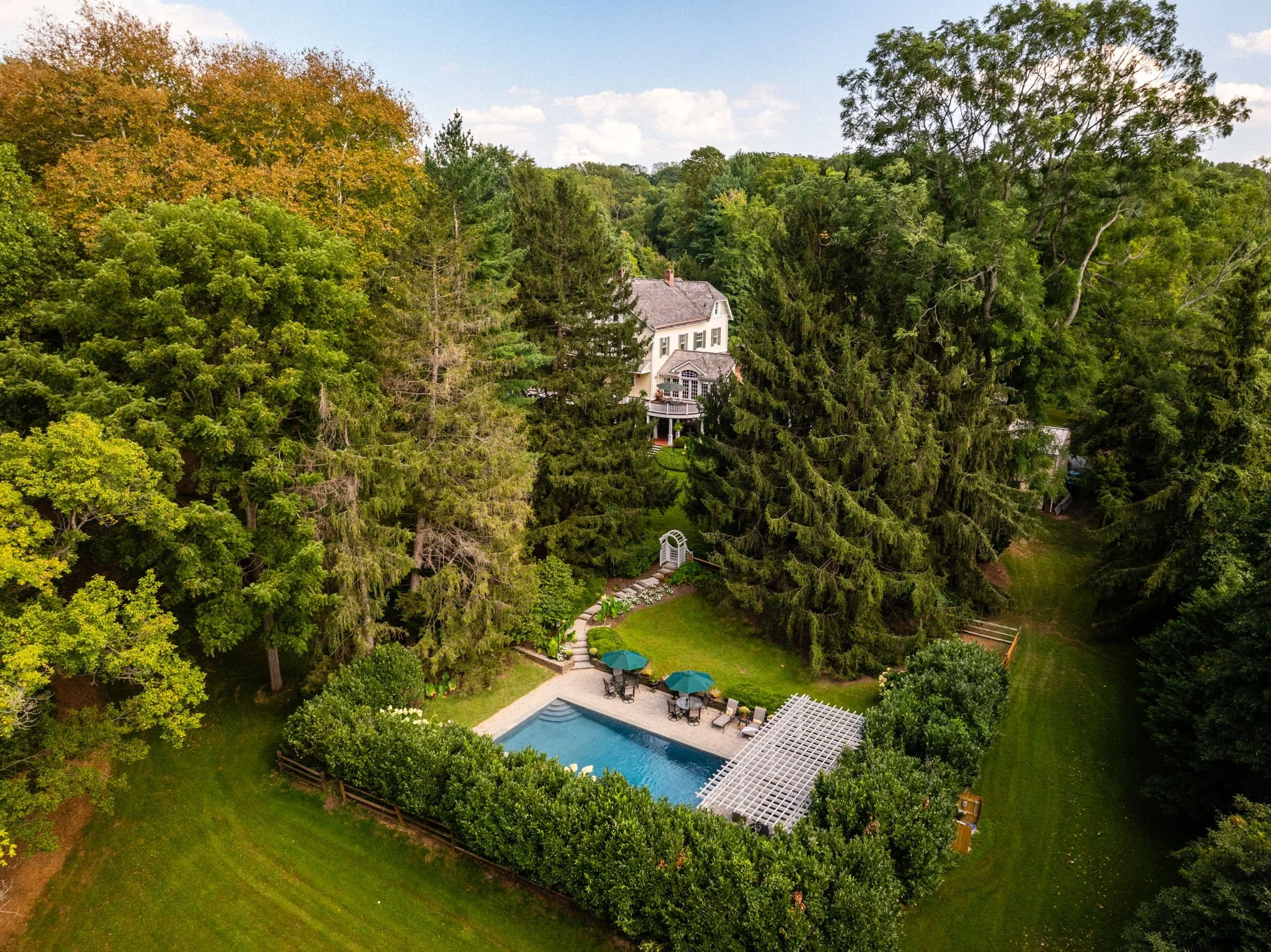 Aerial view of a backyard with a swimming pool, surrounded by trees and a large white house in the background.