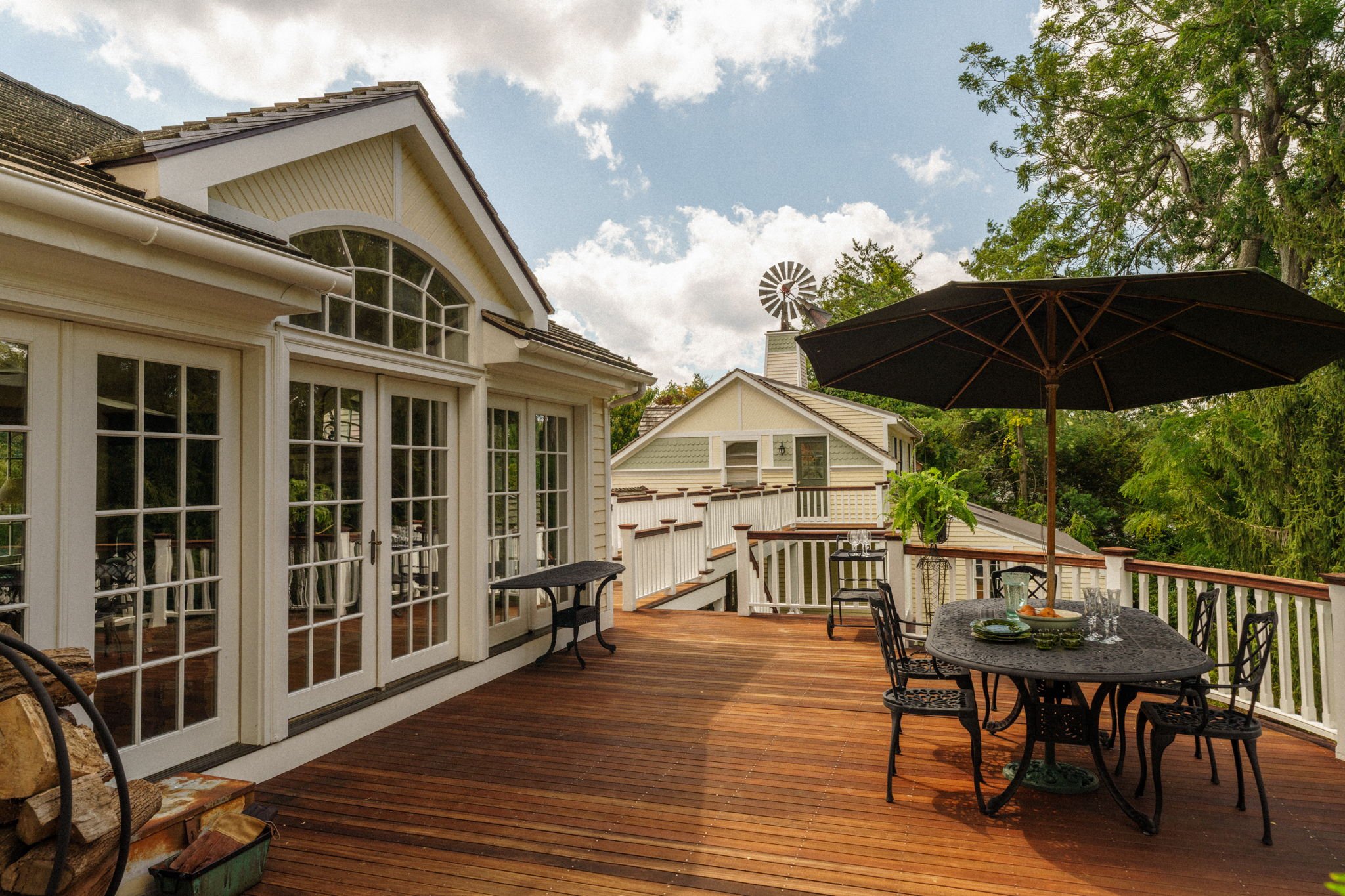 A wooden outdoor deck attached to a house with large glass double doors and a small table with a chair underneath a large black umbrella. There are trees and other houses in the background under a partly cloudy sky.